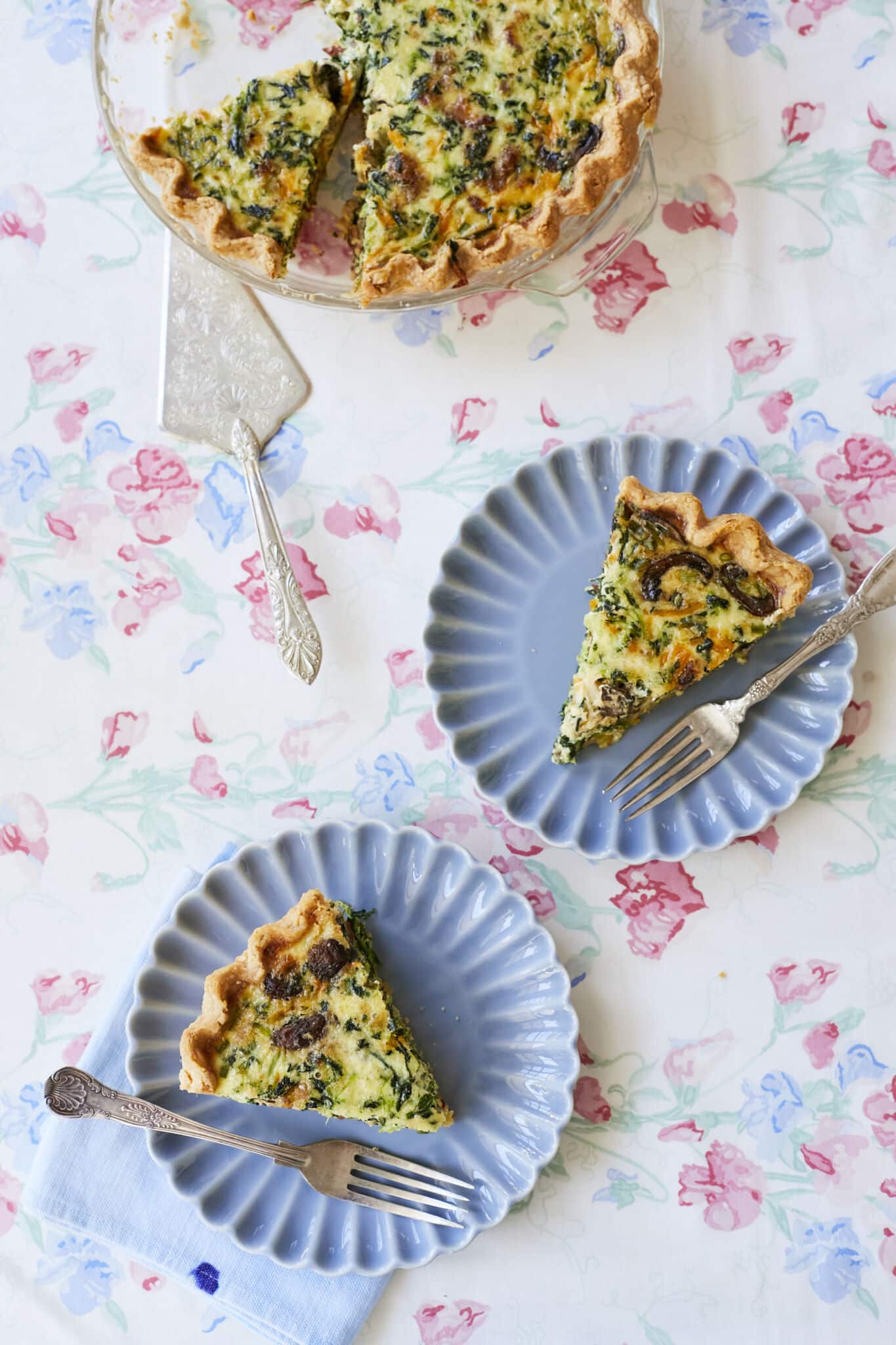 Two slices of homemade quiche are served on blue plates beneath the rest of the quiche, which rests in the baking dish.