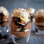 Homemade Chocolate Banana Pudding is served in five individual glasses. The pudding is layered with bananas and crushed Oreo cookies and topped with homemade whipped cream.