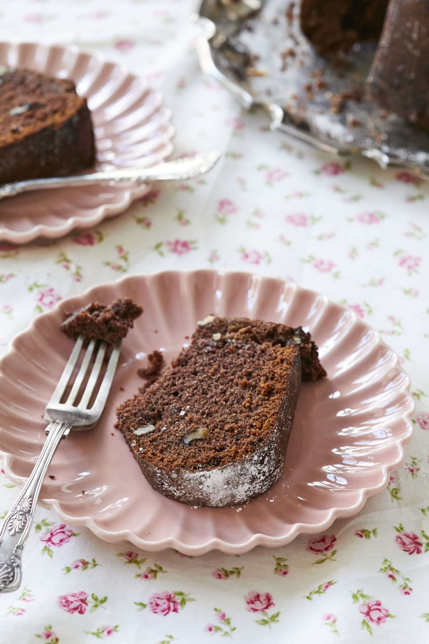 A slice of applesauce cake is served on a pink dish. The cake is brown, colored by cocoa powder, and there are walnuts throughout. 
