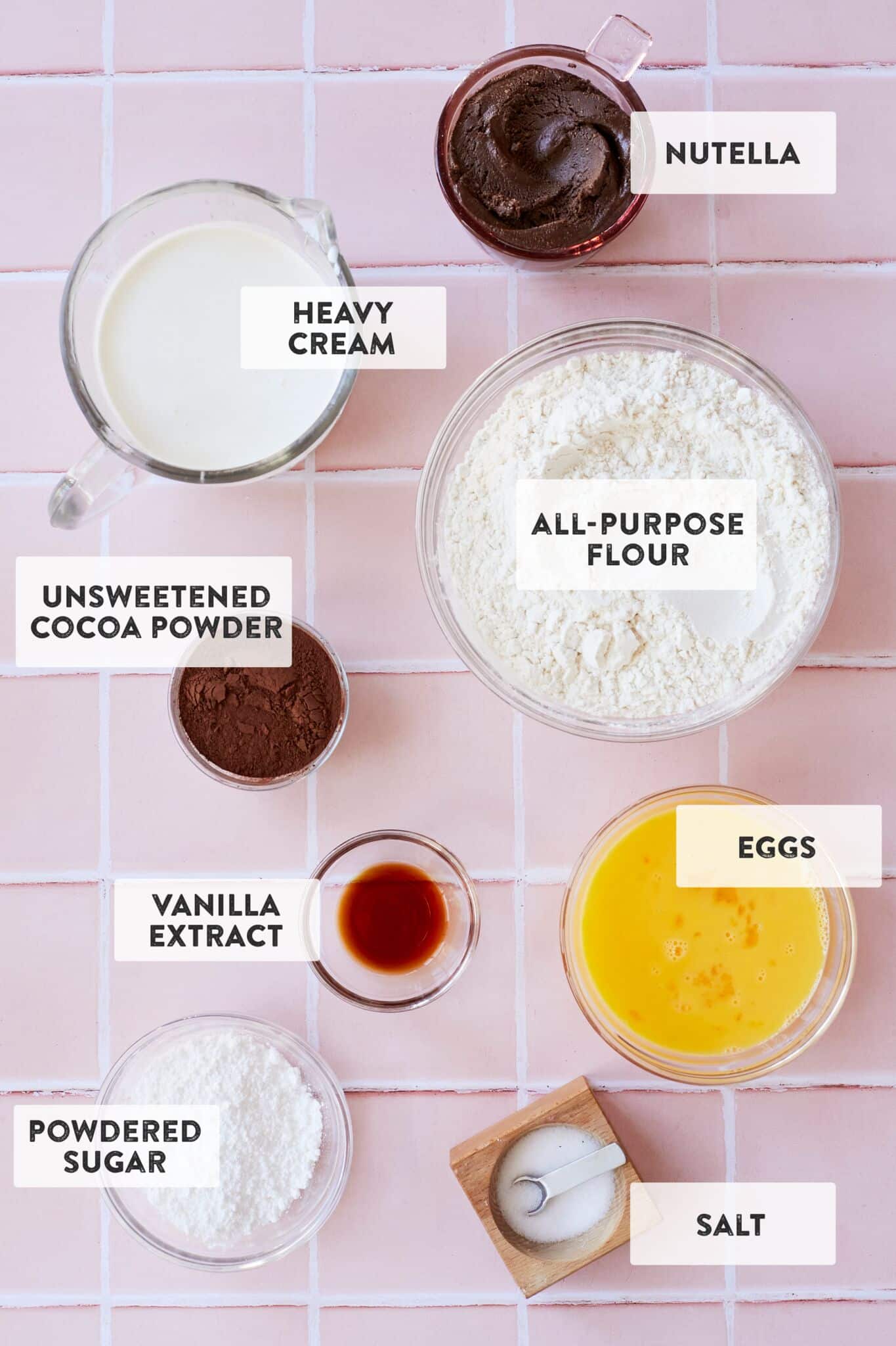 All of the ingredients laid out in preparation bowls: heavy cream, all-purpose flour, cocoa powder, salt, eggs, vanilla extract, powdered sugar, and Nutella, on a pink counter top. 
