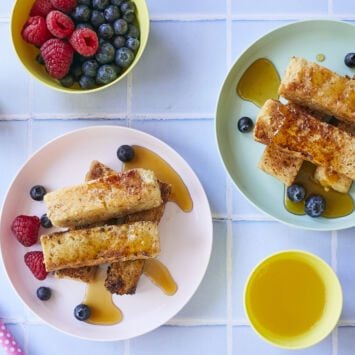 Golden brown French toast sticks paired with Fresh raspberries and blueberries, with syrup on the side.