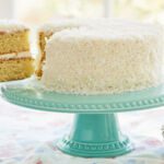 A slice was cut from and being lifted off the perfectly baked two-layer moist coconut cake, with white frosting between layers and on the outside, covered with crunchy coconut.