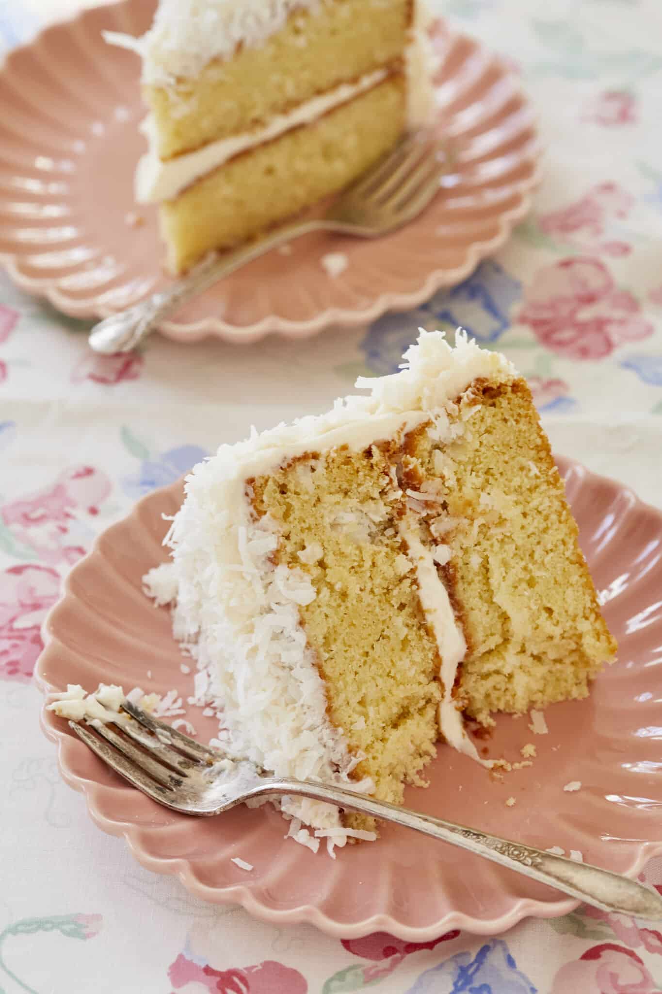 Two slices are served with fork on the plates, showing very moist and soft crumb, smooth frosting and crunchy flakes. . 