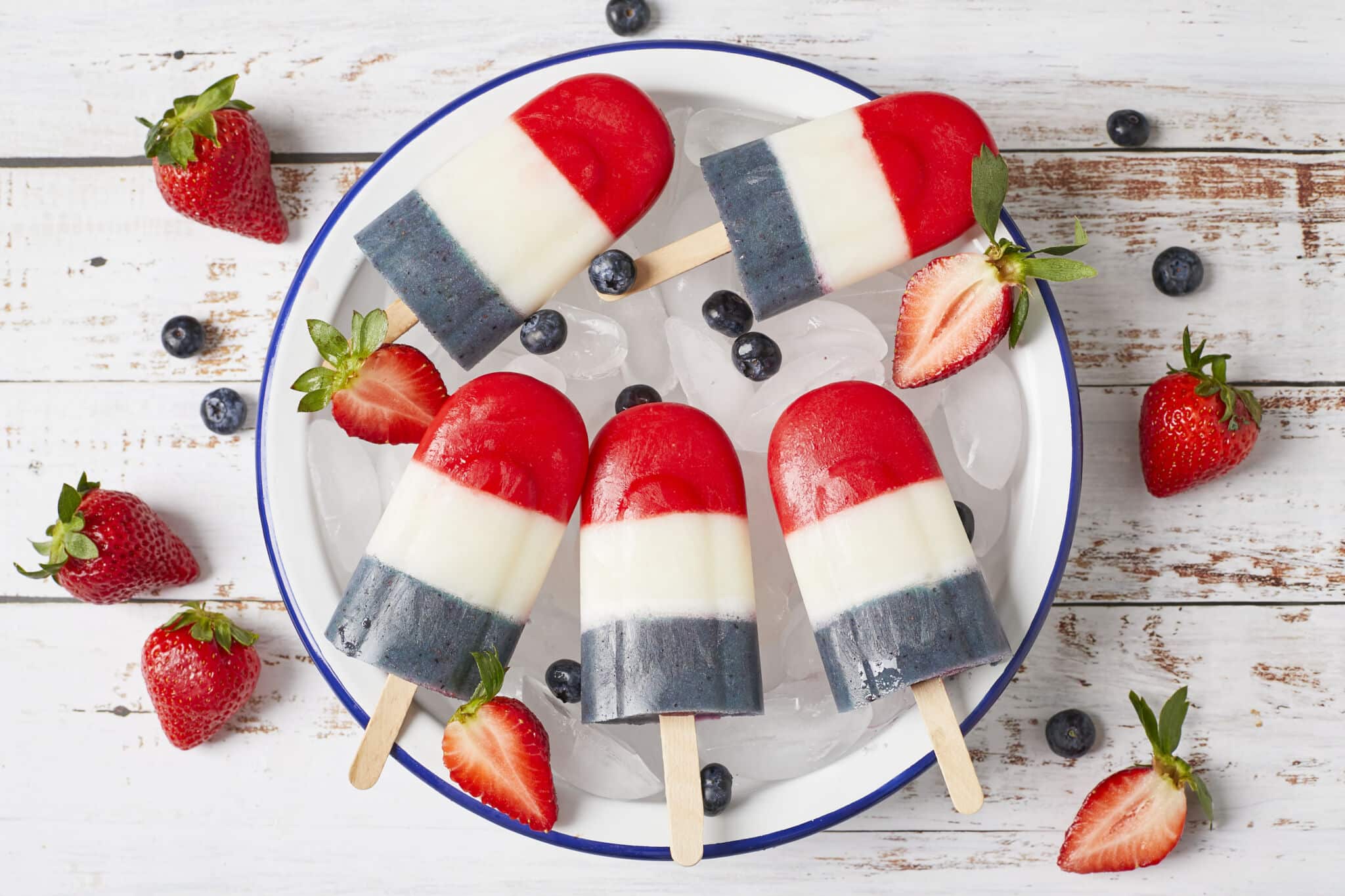 5 Homemade Bomb Pop Popsicles with vibrant red, white and blue colors, served on a round blue-edged white enamel platter with ice cubes. Fresh blueberries and strawberries are on the platter and around the platter.