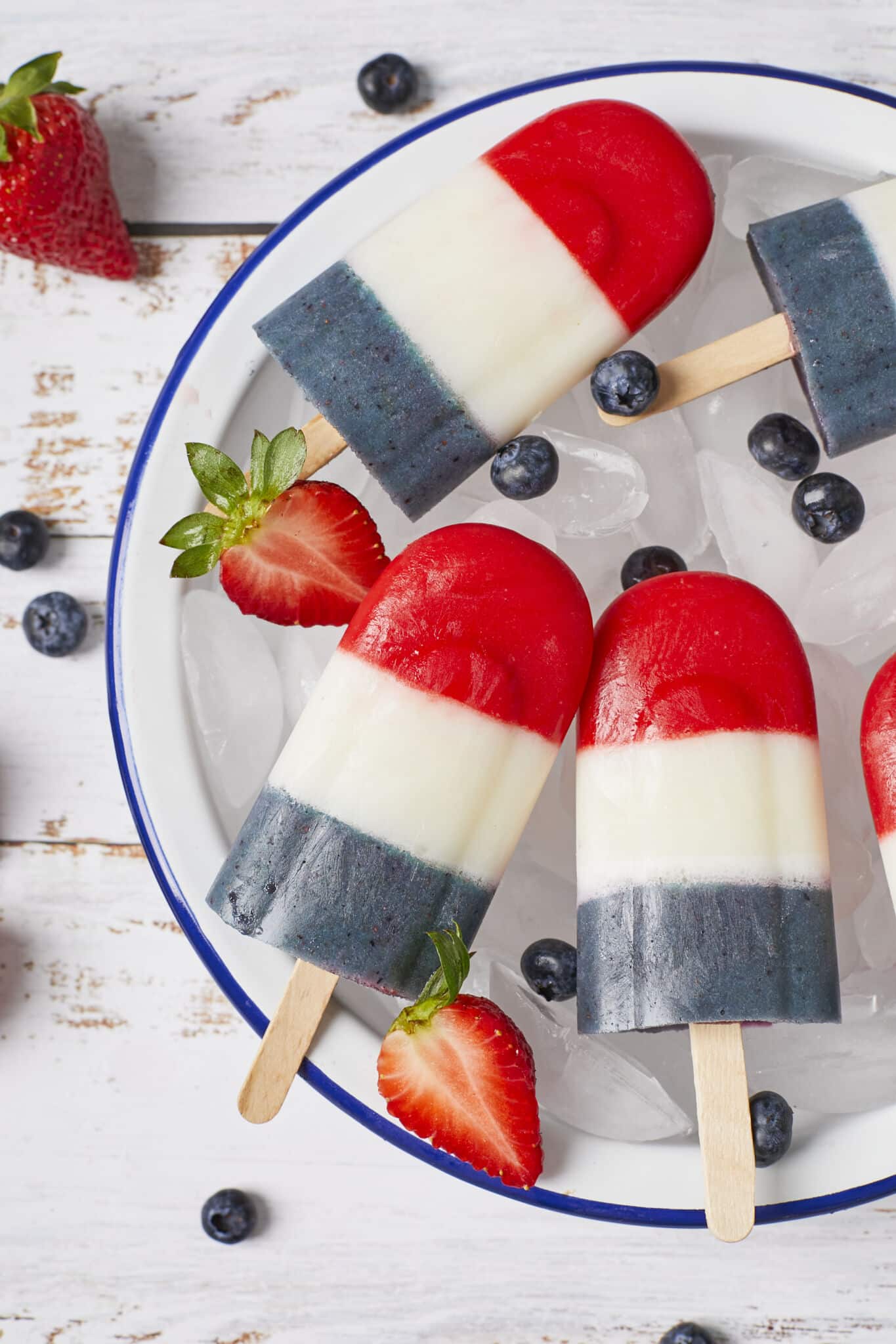 Homemade Bomb Pop Popsicles with vibrant red, white and blue colors, served on a round blue-edged white enamel platter with ice cubes and fresh blueberries. More fresh blueberries and one strawberry are on the side around the platter. 