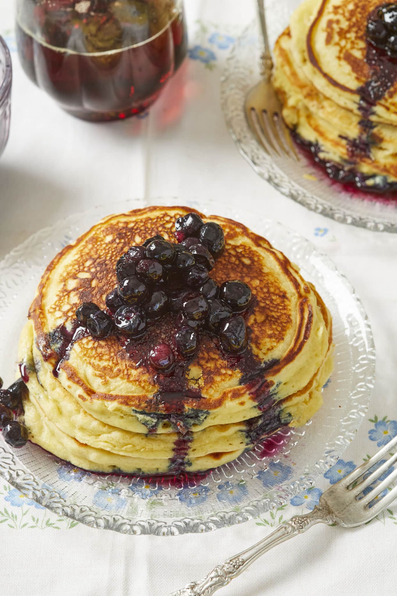 Two stacks of thick, fluffy and soft lemon ricotta pancakes topped with juicy sweet macerated blueberries, served on glass plates and ready to enjoy.