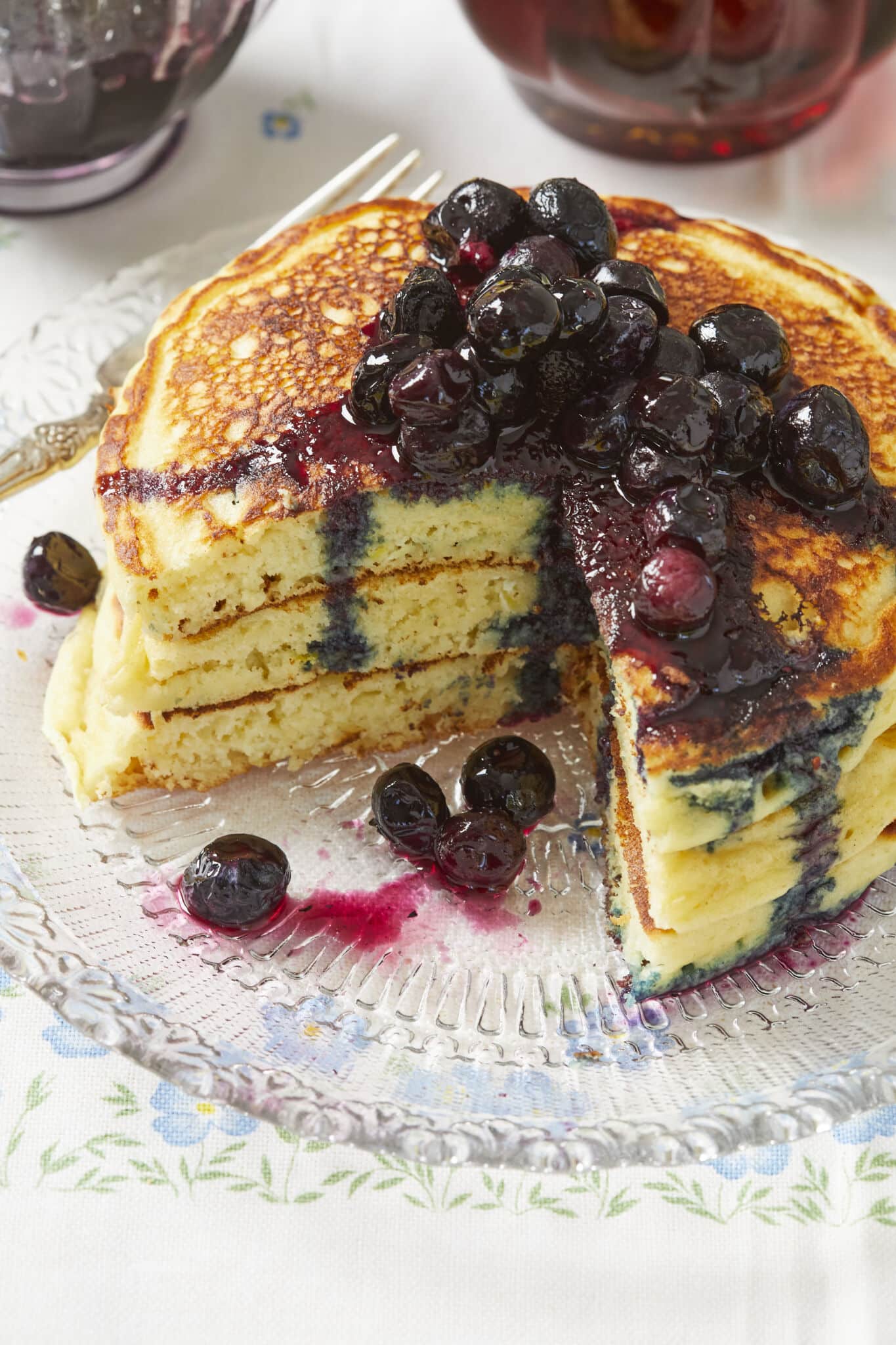A stack of thick, bubbly and fluffy pancakes topped with Macerated Blueberrie are served on a glass place, with a quarter of the stack removed. 