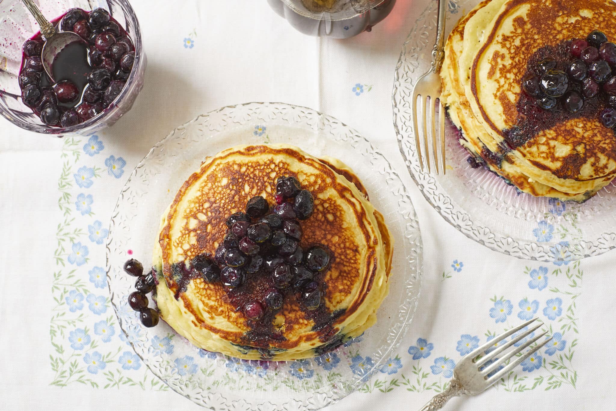 Two stacks of thick, fluffy and soft lemon ricotta pancakes topped with juicy sweet macerated blueberries, served on glass plates and ready to enjoy.