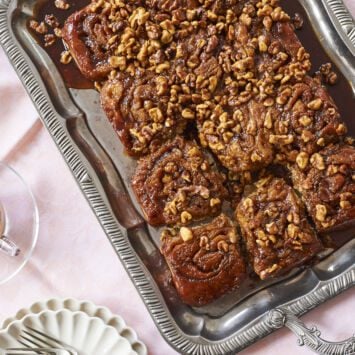 Soft and fluffy cinnamon rolls are packed with bananas and baked golden-brown, served in a silver rectangular plater, topped with toasted walnuts and smooth decadent caramel sauce, perfect to go with tea on the side.