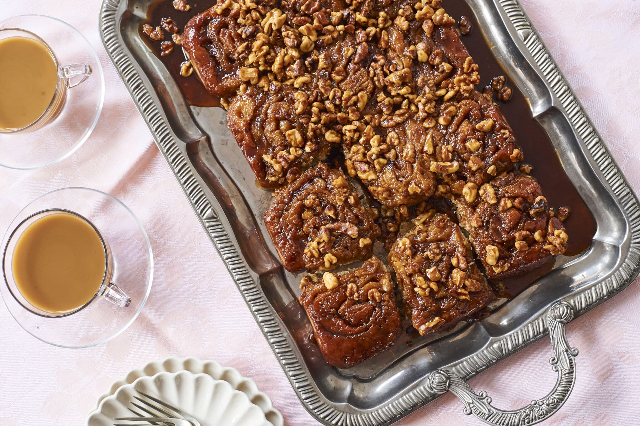 Soft and fluffy cinnamon rolls are packed with bananas and baked golden-brown, served in a silver rectangular plater, topped with toasted walnuts and smooth decadent caramel sauce, perfect to go with tea on the side.