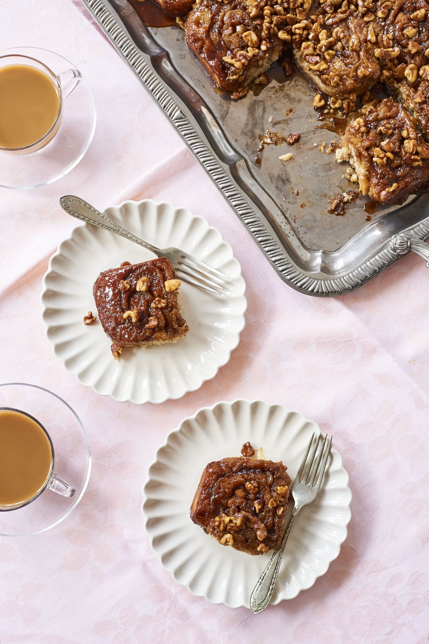 Two of Banana Upside Down Cinnamon Rolls are served on two white scalloped-edge dessert plates with forks, paired with tea. The top is caramelized and crowned with toasted nuts.