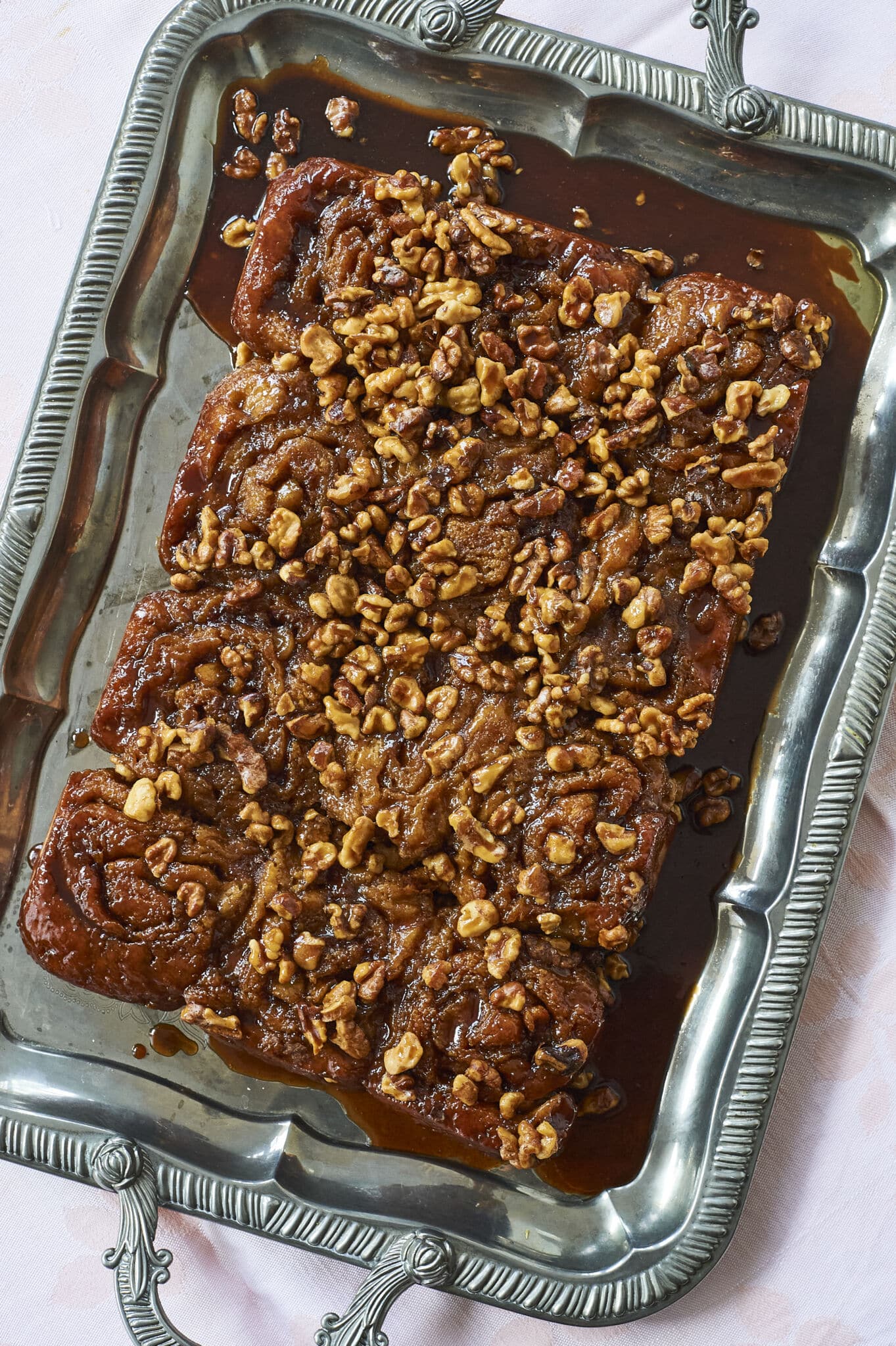 Soft and fluffy cinnamon rolls are packed with bananas and baked golden-brown, served in a silver rectangular plater, topped with toasted walnuts and smooth decadent caramel sauce. 