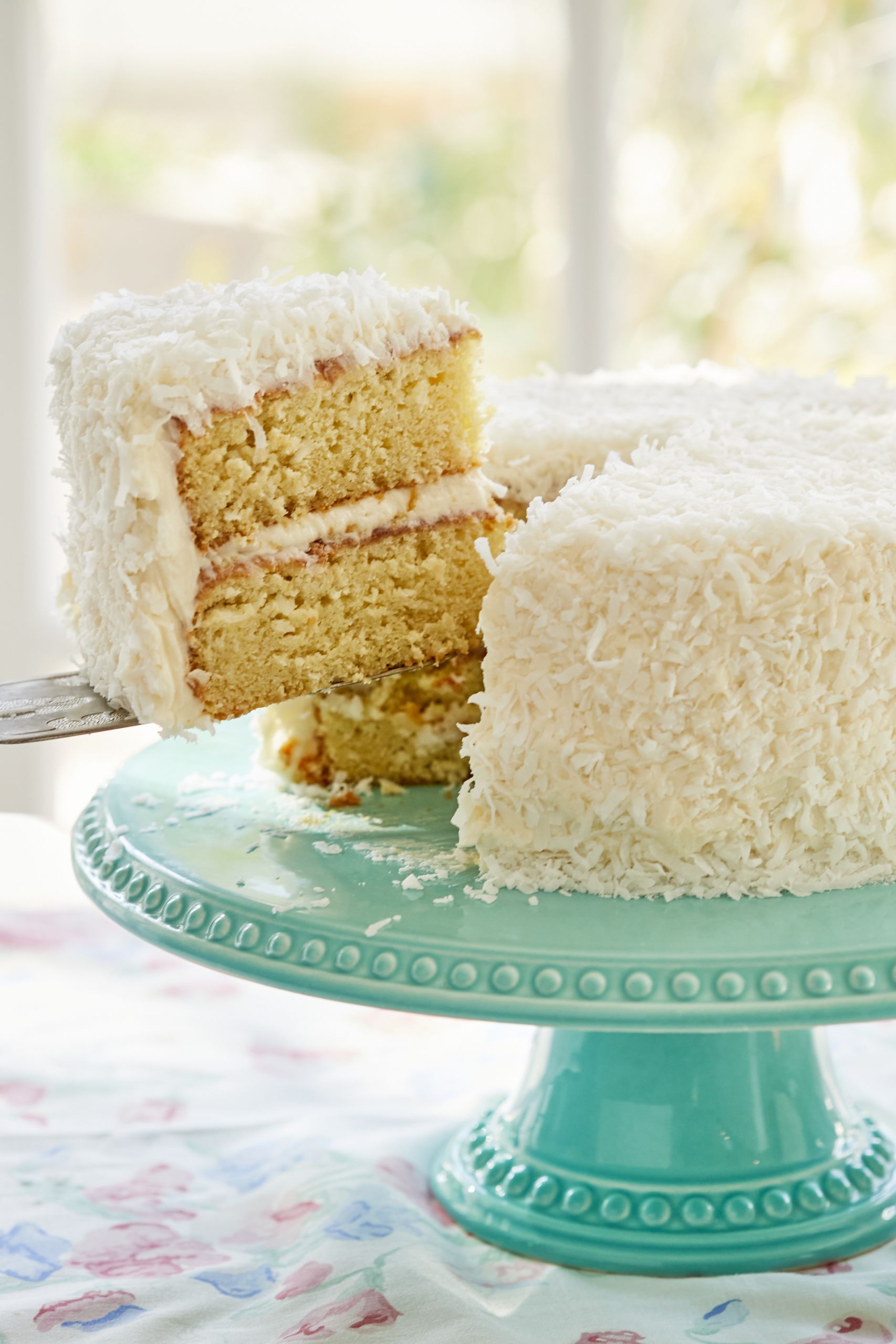 A slice of Coconut Cake is cut from the whole cake. The side shows its moist inside and smooth layer of frosting. 