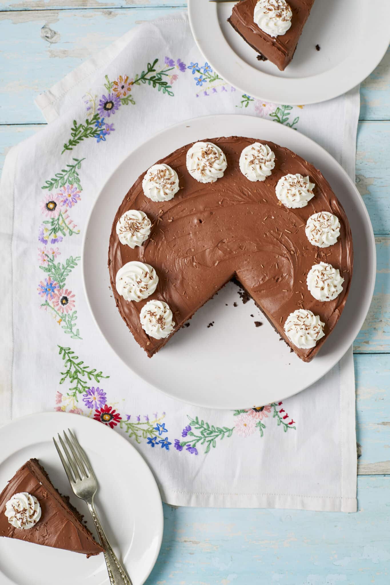 A quarter of the whole no-bake chocolate cheesecake was cut into 2 slices and served on two dessert plates with forks. Each slice has a whipped cream rosette with shaved chocolate. 