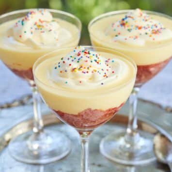 Traditional English Trifle is served in 3 glasses on a silver platter. Each Sherry trifle has a fruity raspberry jelly layer, moist sherry-infused pound cake, velvety creme anglaise, topped with light whipped cream, and colorful classic sprinkles.