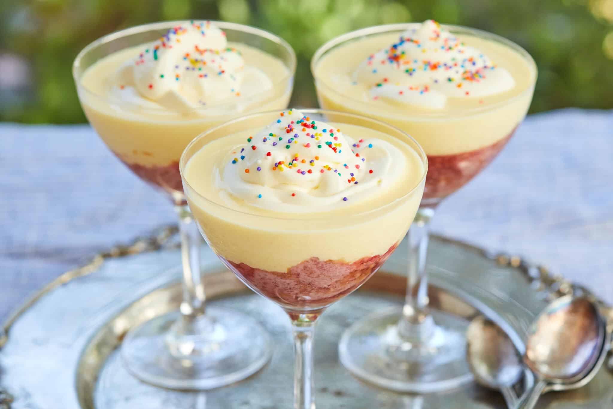 Traditional English Trifle is served in 3 glasses on a silver platter. Each Sherry trifle has a fruity raspberry jelly layer, moist sherry-infused pound cake, velvety creme anglaise, topped with light whipped cream, and colorful classic sprinkles.