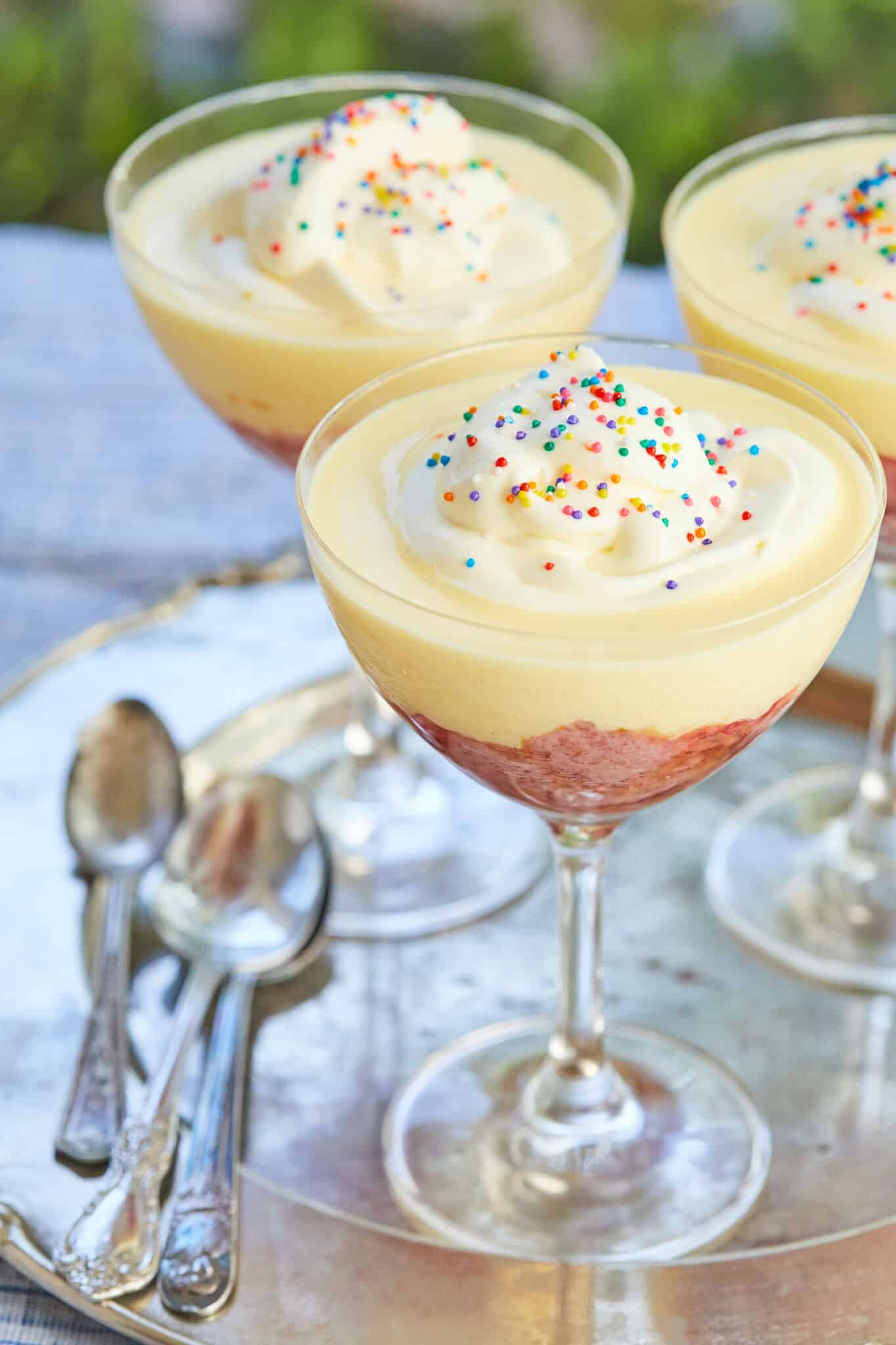 Traditional English Trifle is served in 3 glasses on a silver platter. Each Sherry trifle has a pink fruit-flavored jelly layer, boozy moist sherry infused pound cake, velvety creme anglaise, topped with light whipped cream, and colorful classic sprinkles. 