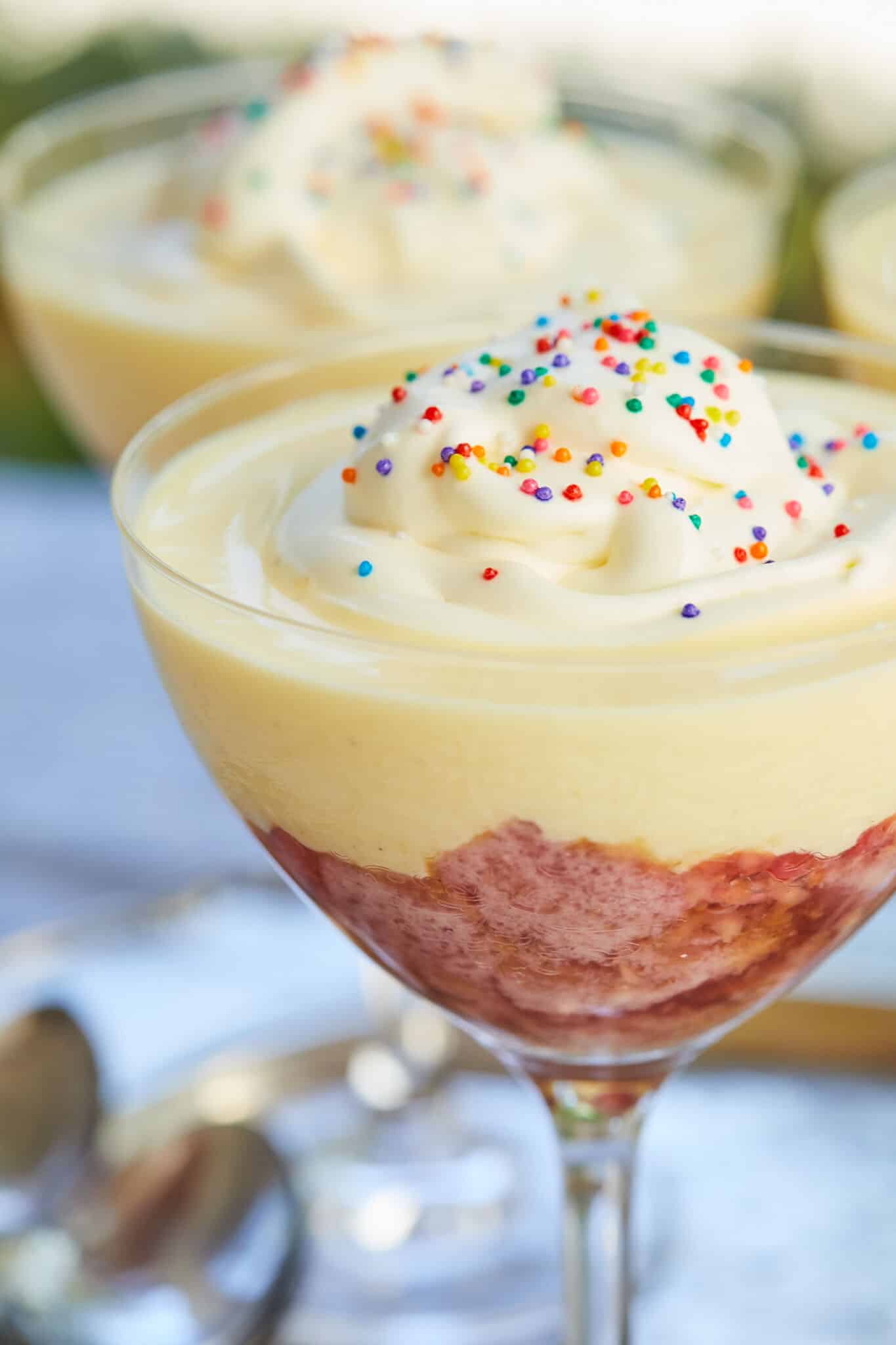 A closes shot of one glass of Sherry trifle with pink sherry soaked moist pound cake and raspberry flavored jelly at the bottom, smooth creamy custard in the middle, and topped with softly whipped cream and colorful sprinkles. 