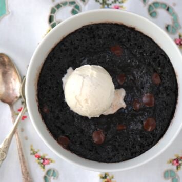 The luscious perfectly microwave-steamed brownie bowl looks fudgy and moist, with an almost black color from dutch-processed cocoa powder, brown color from soft melted chocolate chips and milk color from the smooth velvety vanilla ice cream. Served in a white bowl with a spoon and a fork on the left.