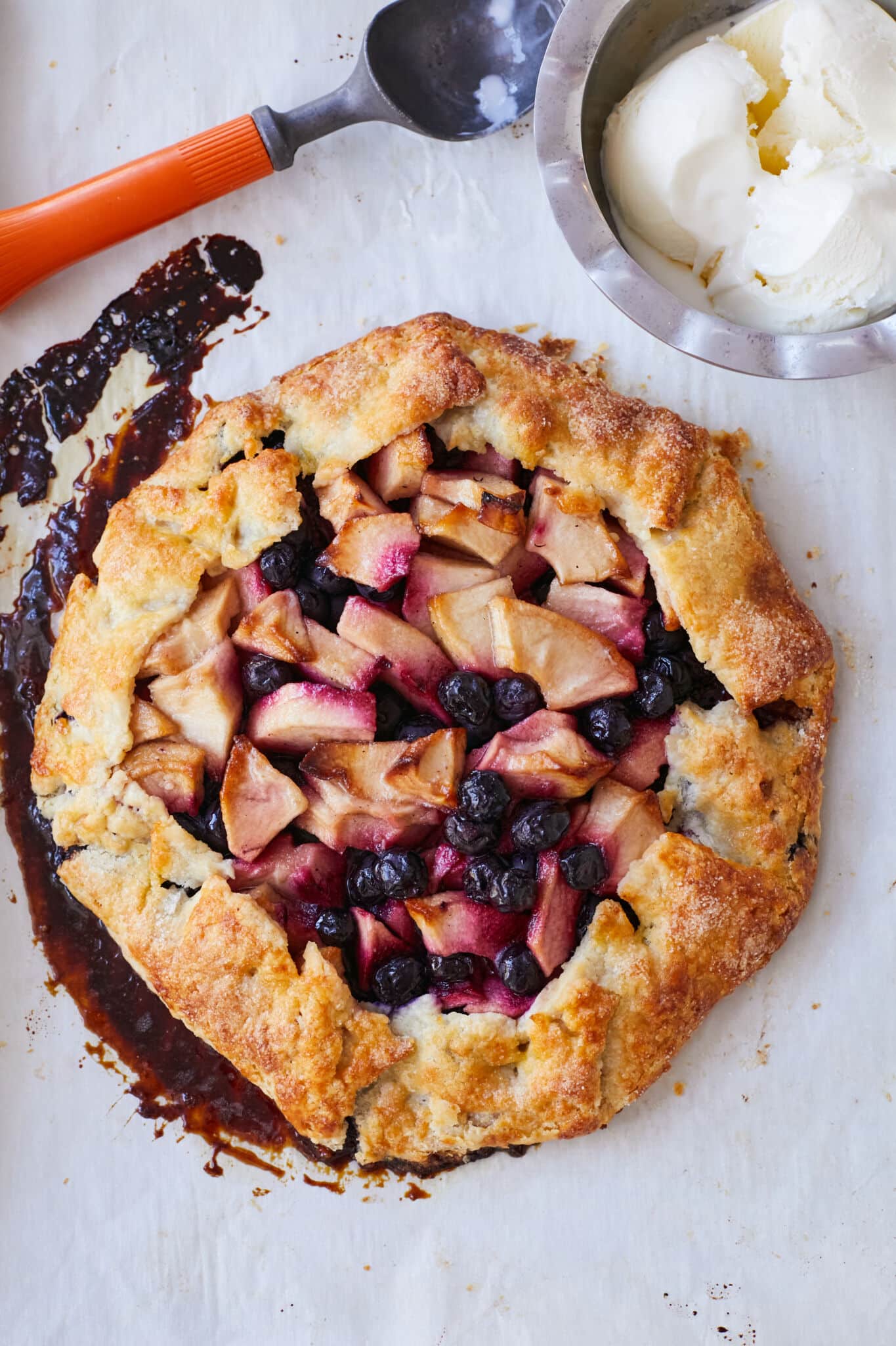 An Apple Blueberry and Vanilla Bean Galette is baked golden brown on the edges with juicy bubbling apples and blueberries in the center. It's paired with a bowl of vanilla ice cream in the top right corner. 
