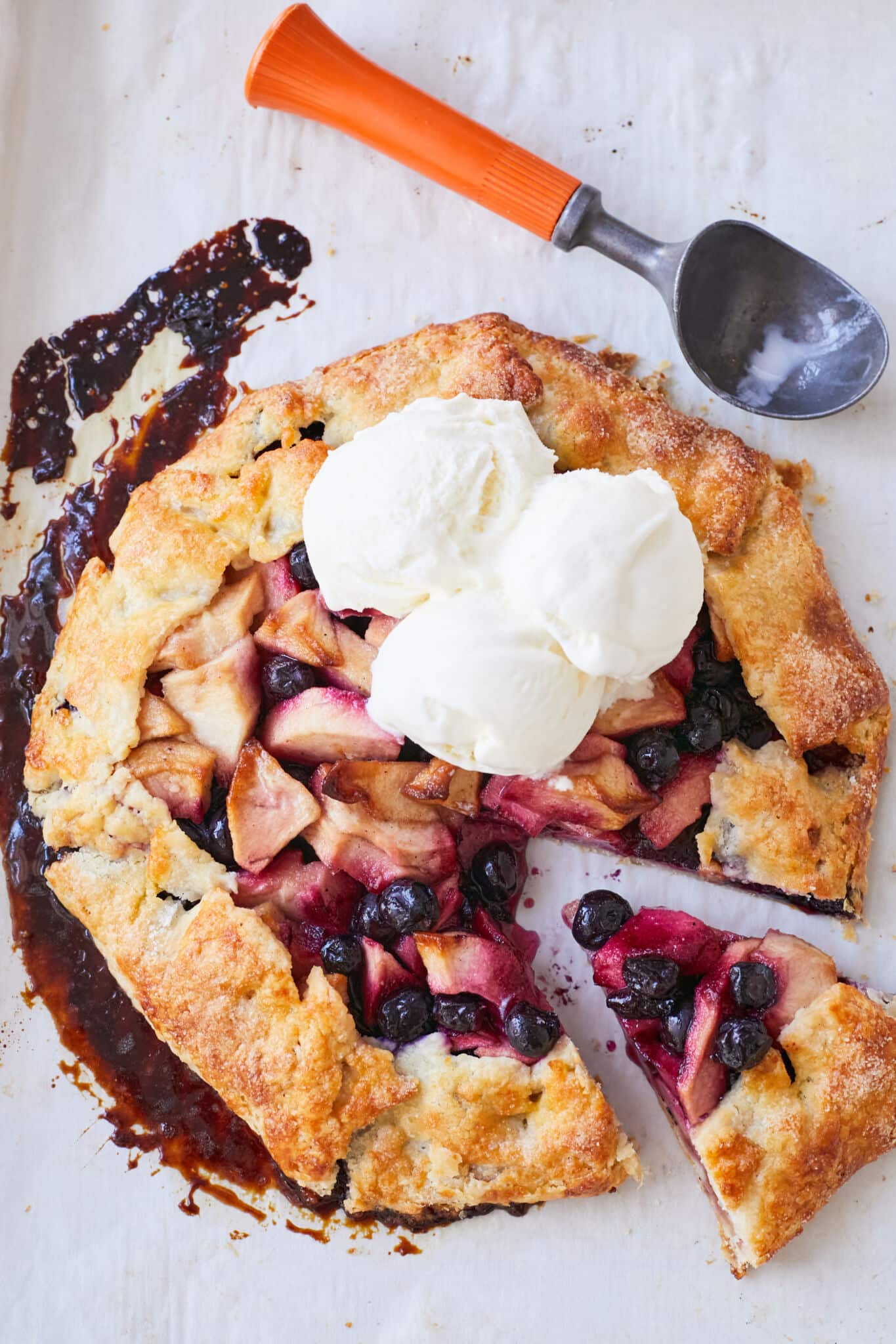 An Apple Blueberry and Vanilla Bean Galette is baked golden brown on the edges with juicy bubbling apples and blueberries in the center. It's topped with 3 big scoops of vanilla ice cream. A piece was sliced off the whole galette, ready for serving. 