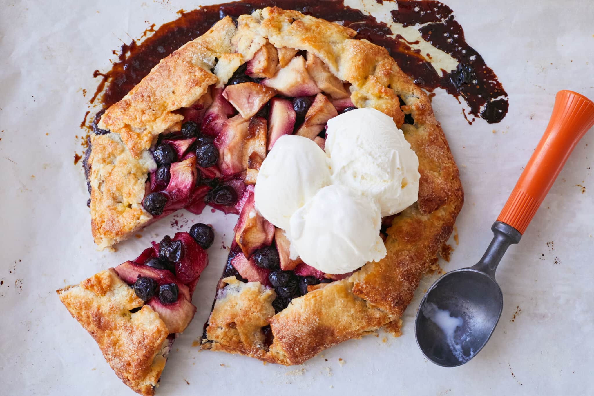 An Apple Blueberry and Vanilla Bean Galette is baked golden brown on the edges with juicy bubbling apples and blueberries in the center. It's served with 3 big scoops of vanilla ice cream on top. A serving was sliced of the whole galette.