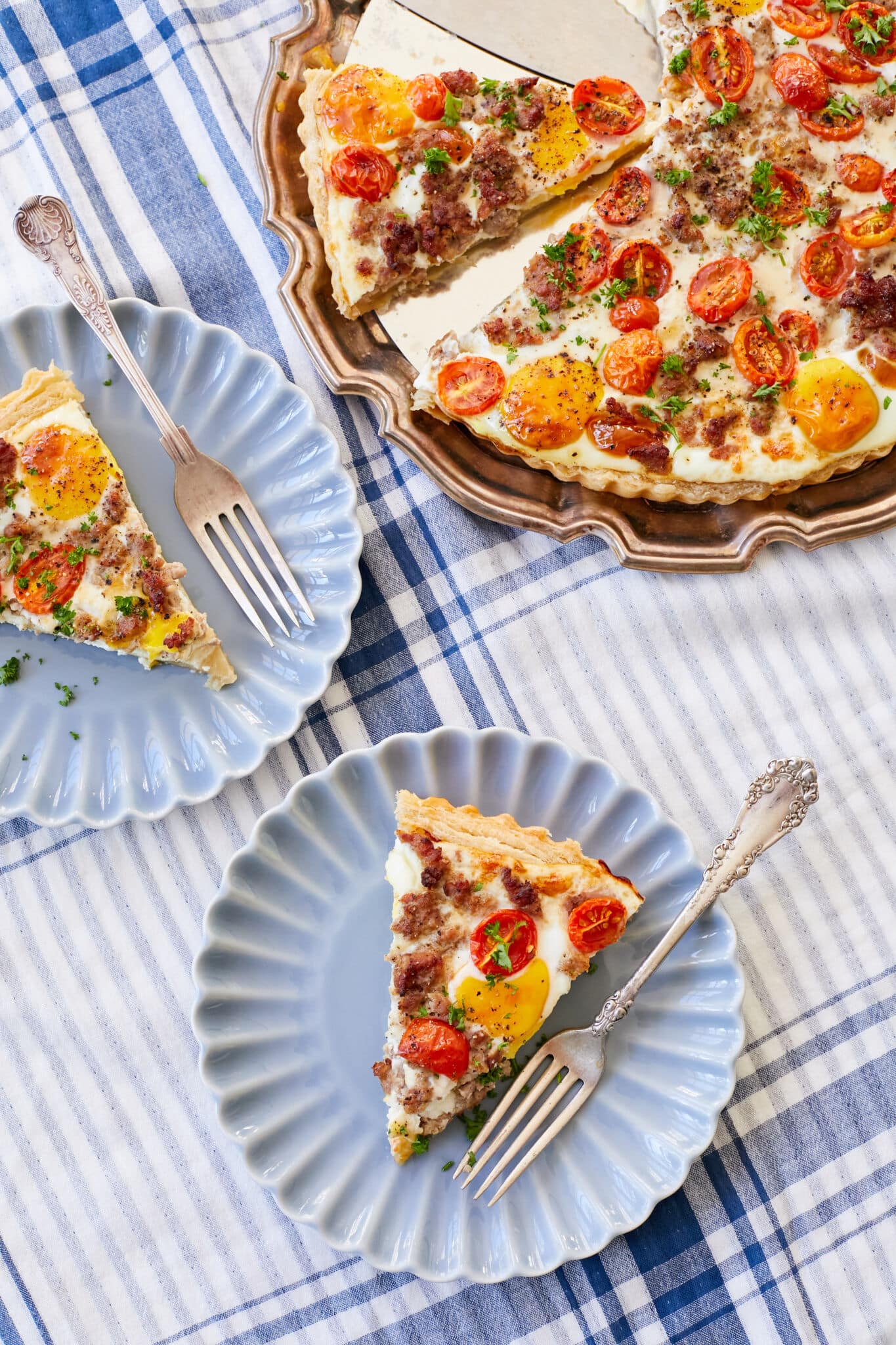 The Complete Breakfast Tart is placed and divided in a rose-gold platter. Two slices are served on two blue scalloped-edge plates with a fork on each. The tart has golden crust and is loaded with savory sausage, melted creamy cheese, bright red cherry tomatoes, eggs and topped with parsley.