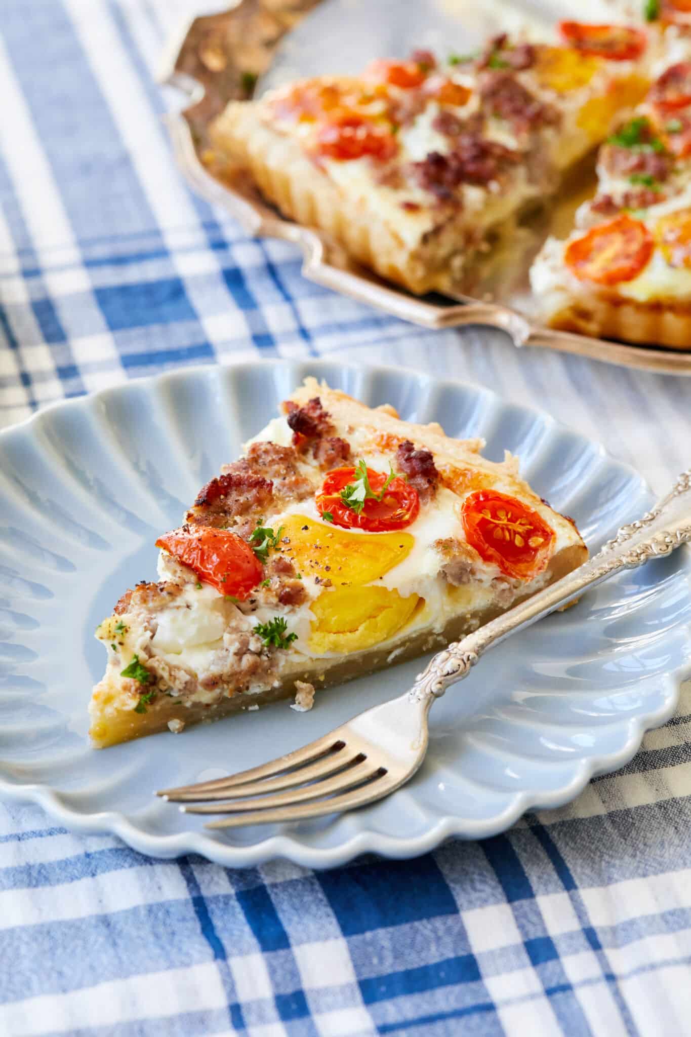 A close shot at a single slice of the Complete Breakfast Tart on a small blue scalloped-edge plate with a fork, showing the golden crust, savory sausage, melted creamy cheese, bright red cherry tomatoes, eggs and parsley. The rest of the tart is in a rose-gold platter in the upper right corner of the image. 