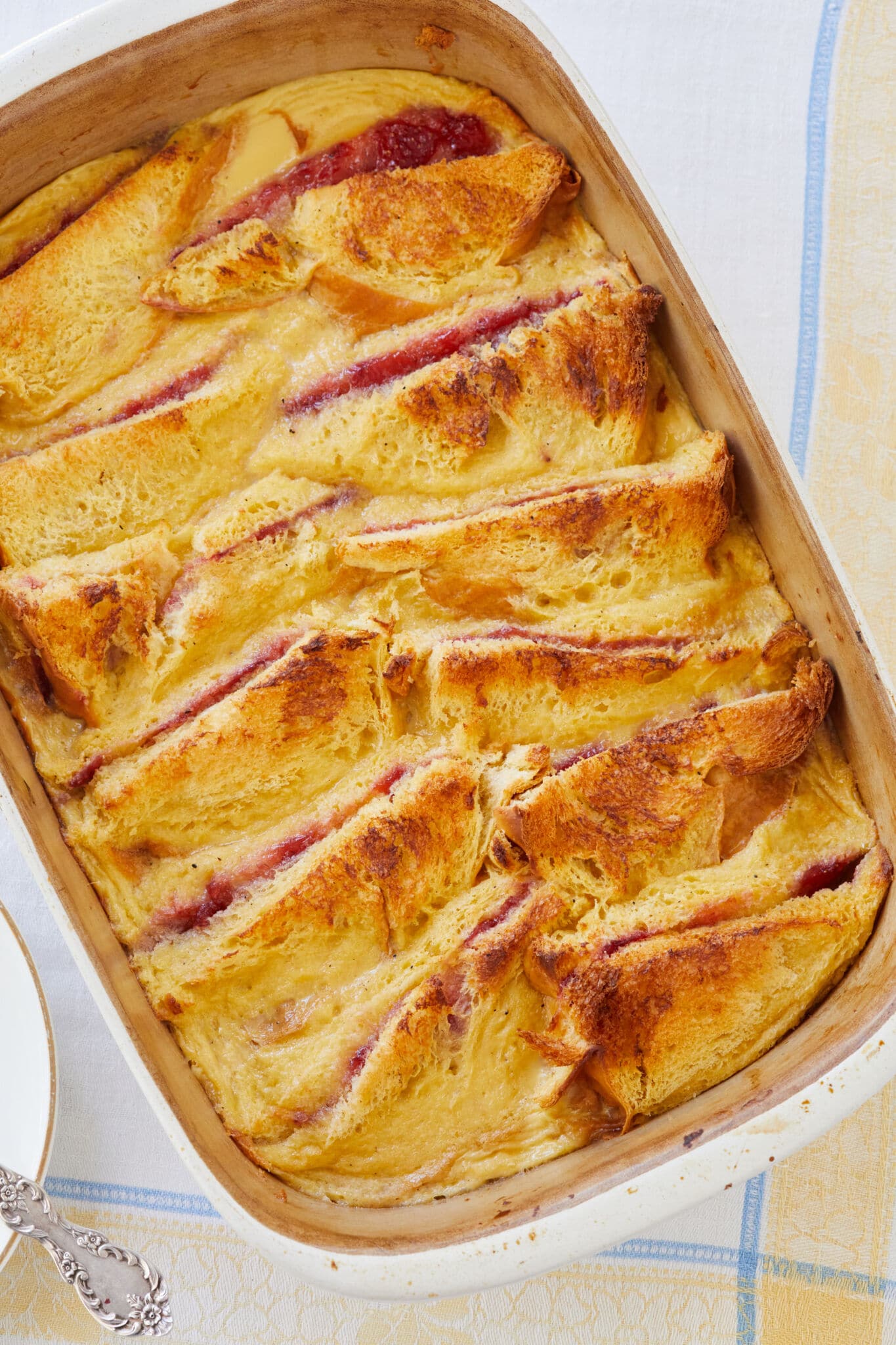 Butter and Jam Bread Pudding is baked to perfection in the whited baking dish. It has a golden-brown and slightly crispy top, glistening with melted butter from the base and vibrant red strawberry jam between slices of the brioche. The rich velvety custard is visible around edges.