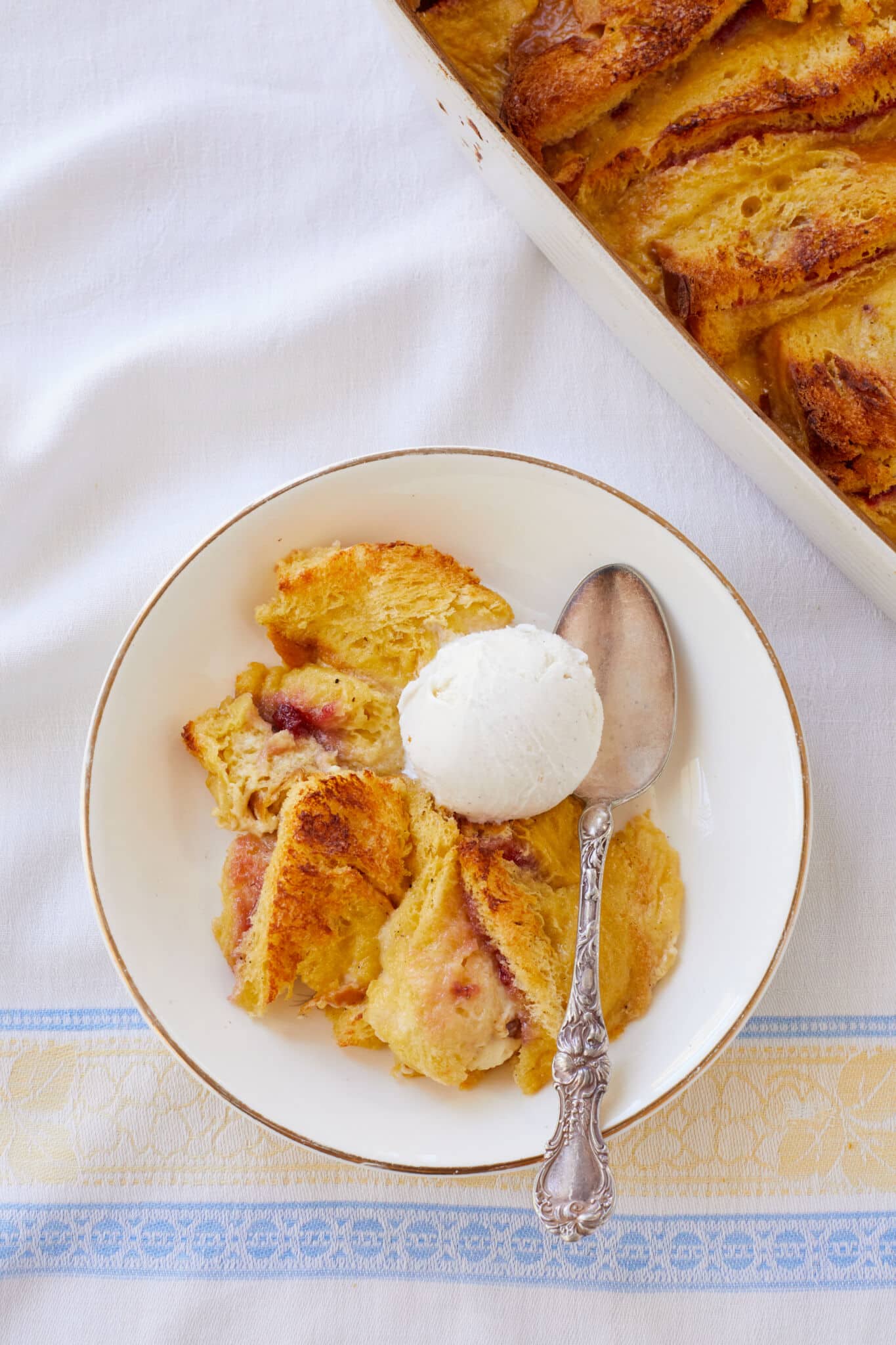 Butter and Jam Bread Pudding is served in a golden-edge white ceramic dessert bowl with a scoop of ice cream on top and a silver spoon. The bread is baked golden-brown and slightly crispy on top, glistening with melted butter and vibrant red strawberry jam. The rich velvety custard is visible through the brioche's edges. The rest is in the white baking dish in the top right corner of the image. 