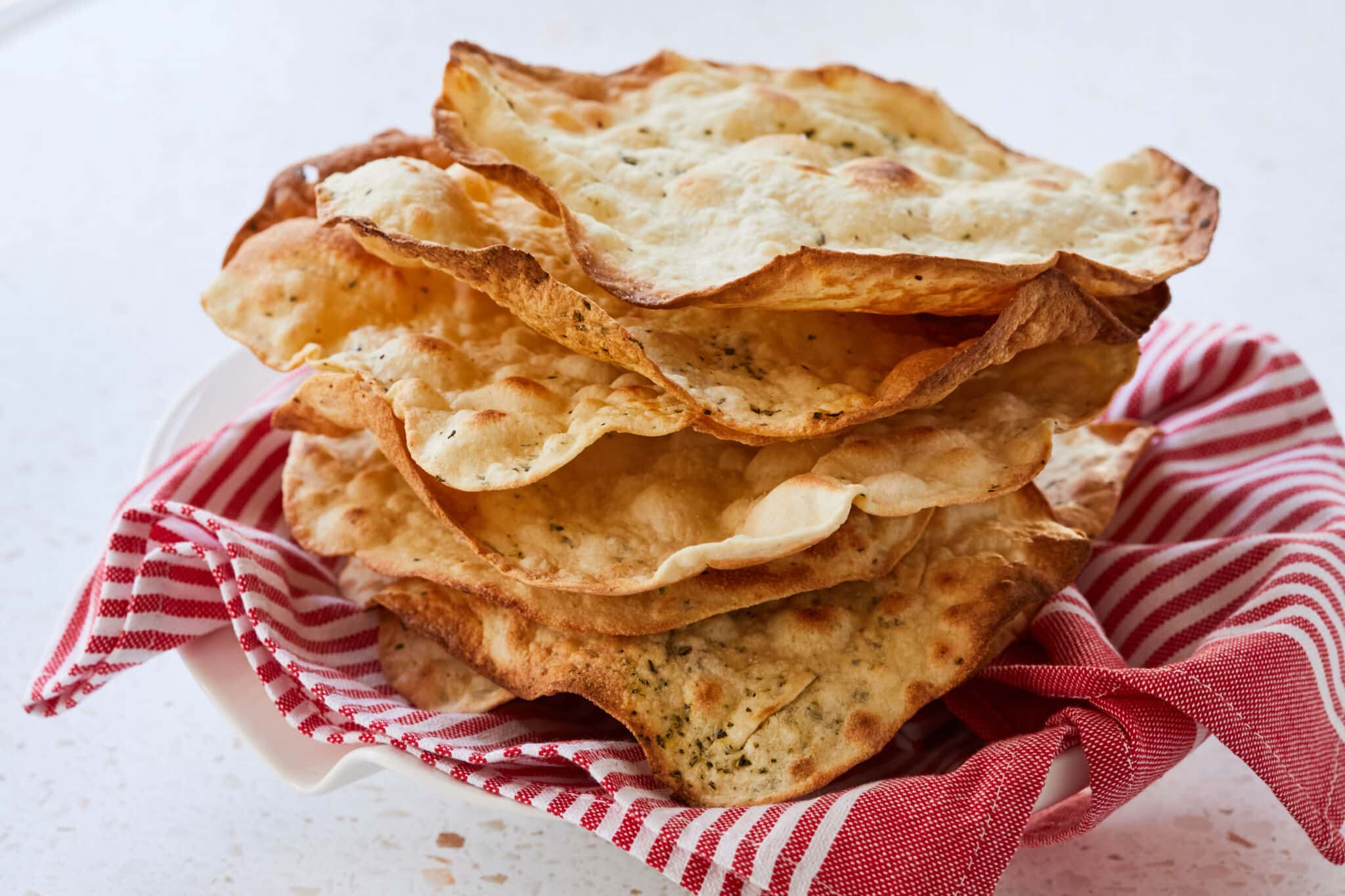 A stack of thin crispy Carta Di Musica is baked to golden brown perfection, sprinkled with flaky sea salt and herbs. Served on a white-red strip towel in a big white platter.