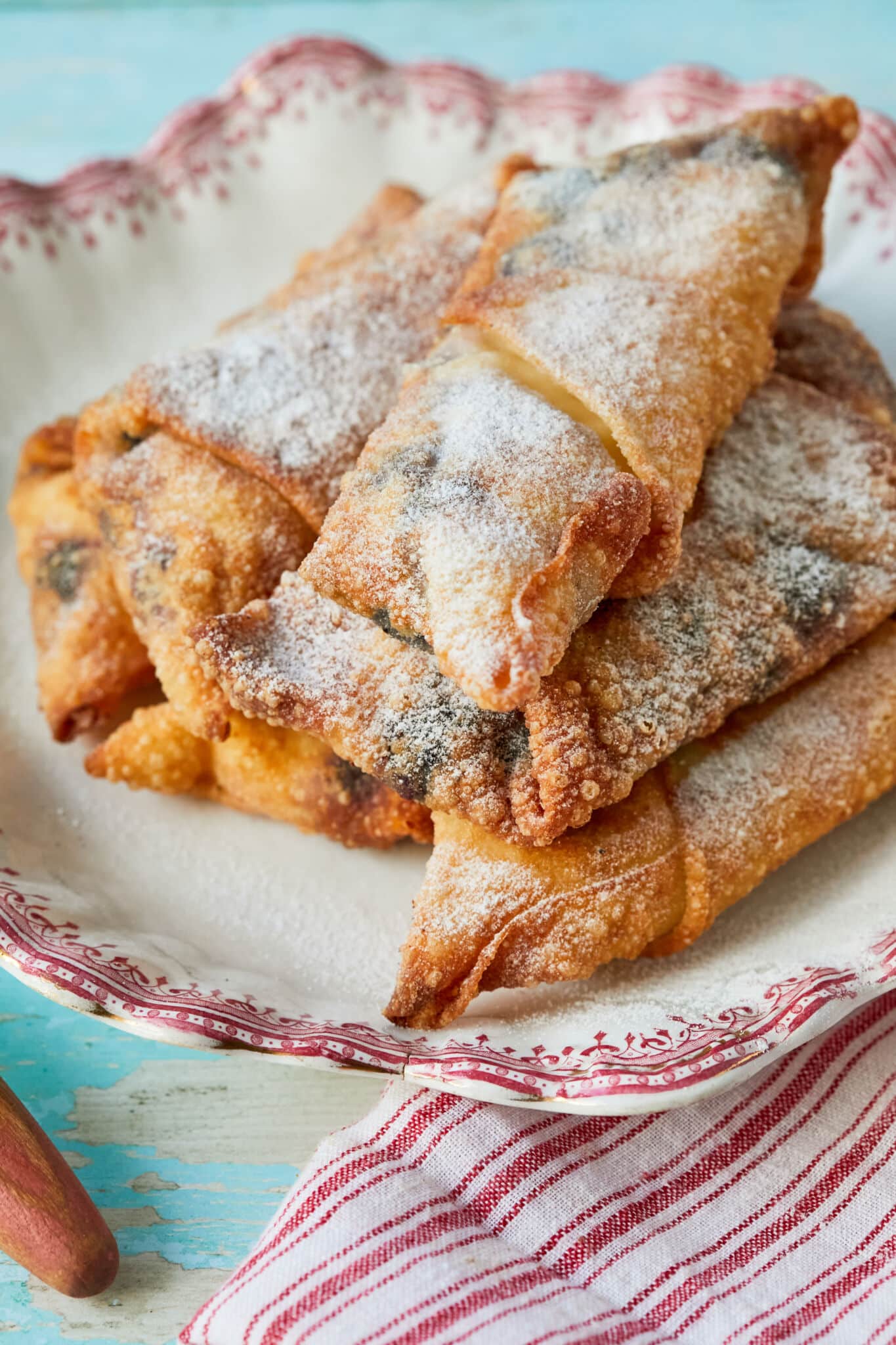Golden crispy Blueberry Cheesecake Egg Rolls are dusted with powdered sugar, served on a red-edge platter. 
