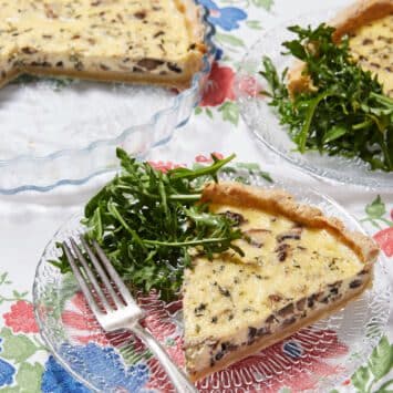 Two slices of Mushroom and Thyme Leaf Tart are cut from the whole tart and served on clear glass plates with forks. The tart has golden, buttery and crumbly crust loaded with velvety custard filling and mushrooms, paired with green earthy thyme leaves. The rest of the tart is in the tart dish.