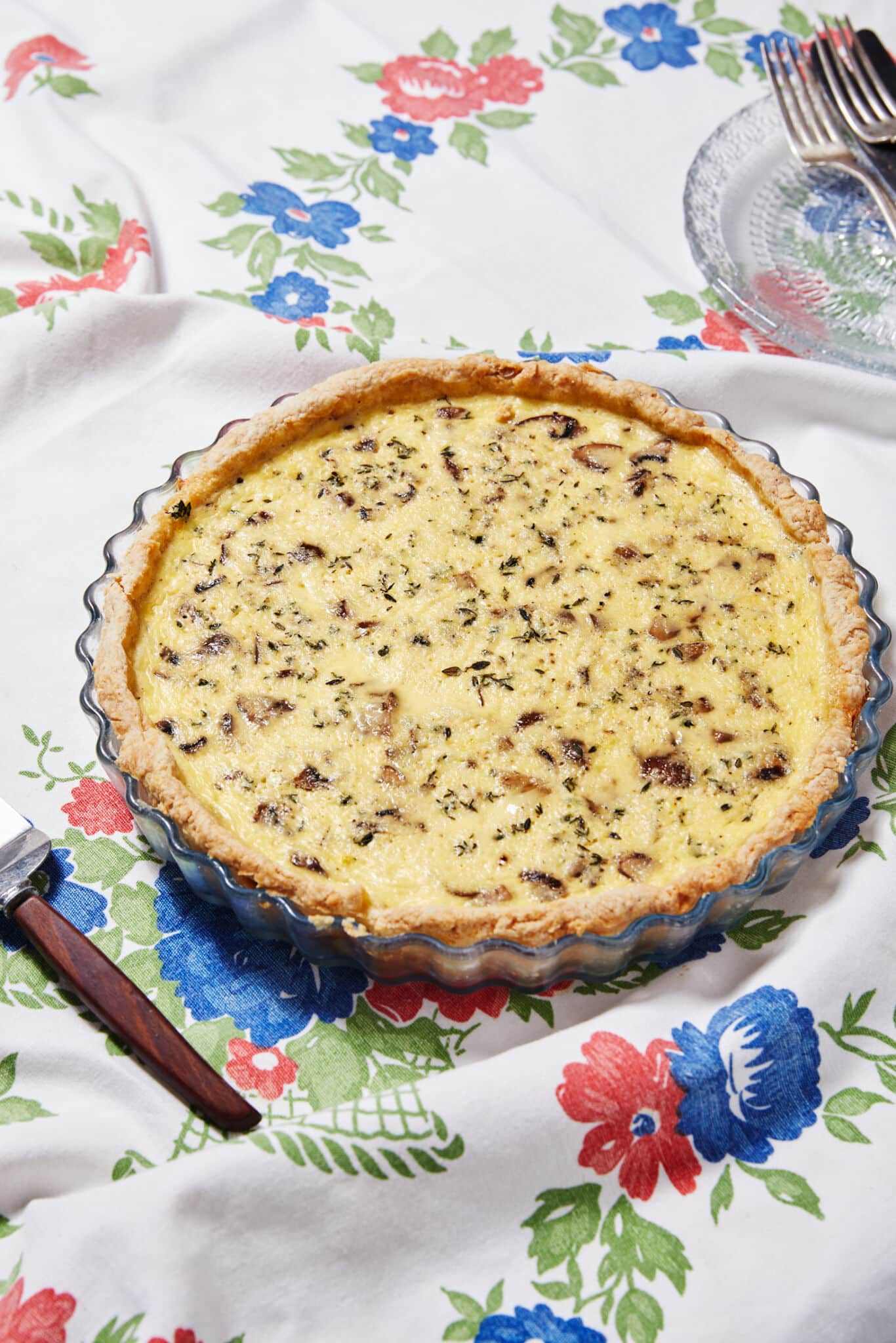 The whole Mushroom and Thyme Leaf Tart is baked in a glass tart dish, with golden, buttery and crumbly crust loaded with velvety custard filling and mushrooms. A tart server, small plate and forks are on the side.