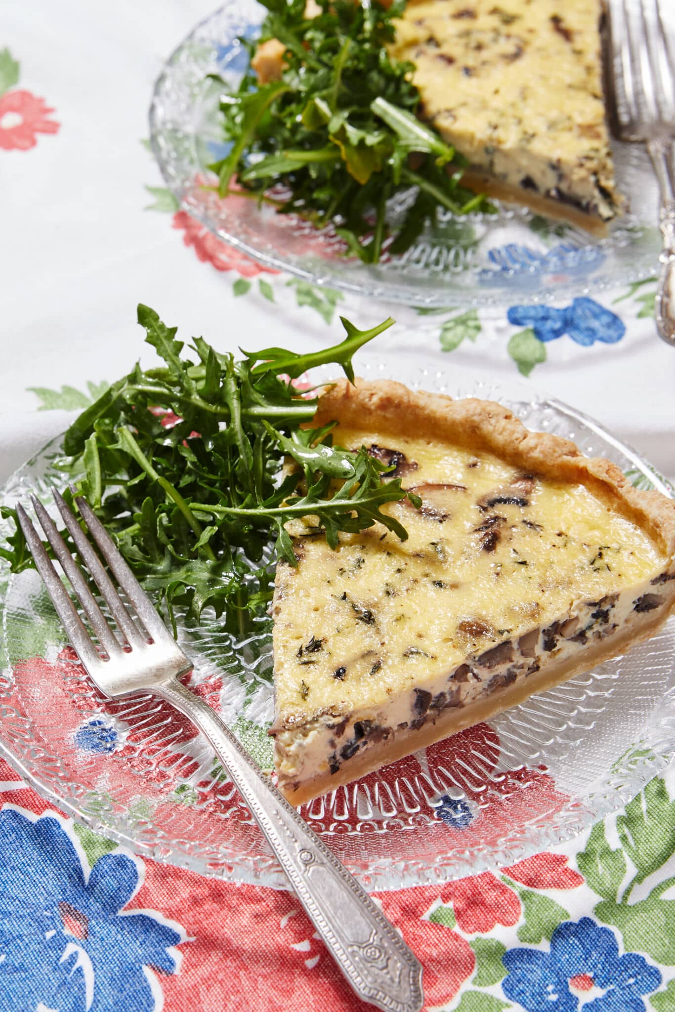 Two slices of Mushroom and Thyme Leaf Tart are served on clear glass plates with forks. The tart has golden, buttery and crumbly crust loaded with velvety custard filling and mushrooms, paired with green earthy thyme leaves.