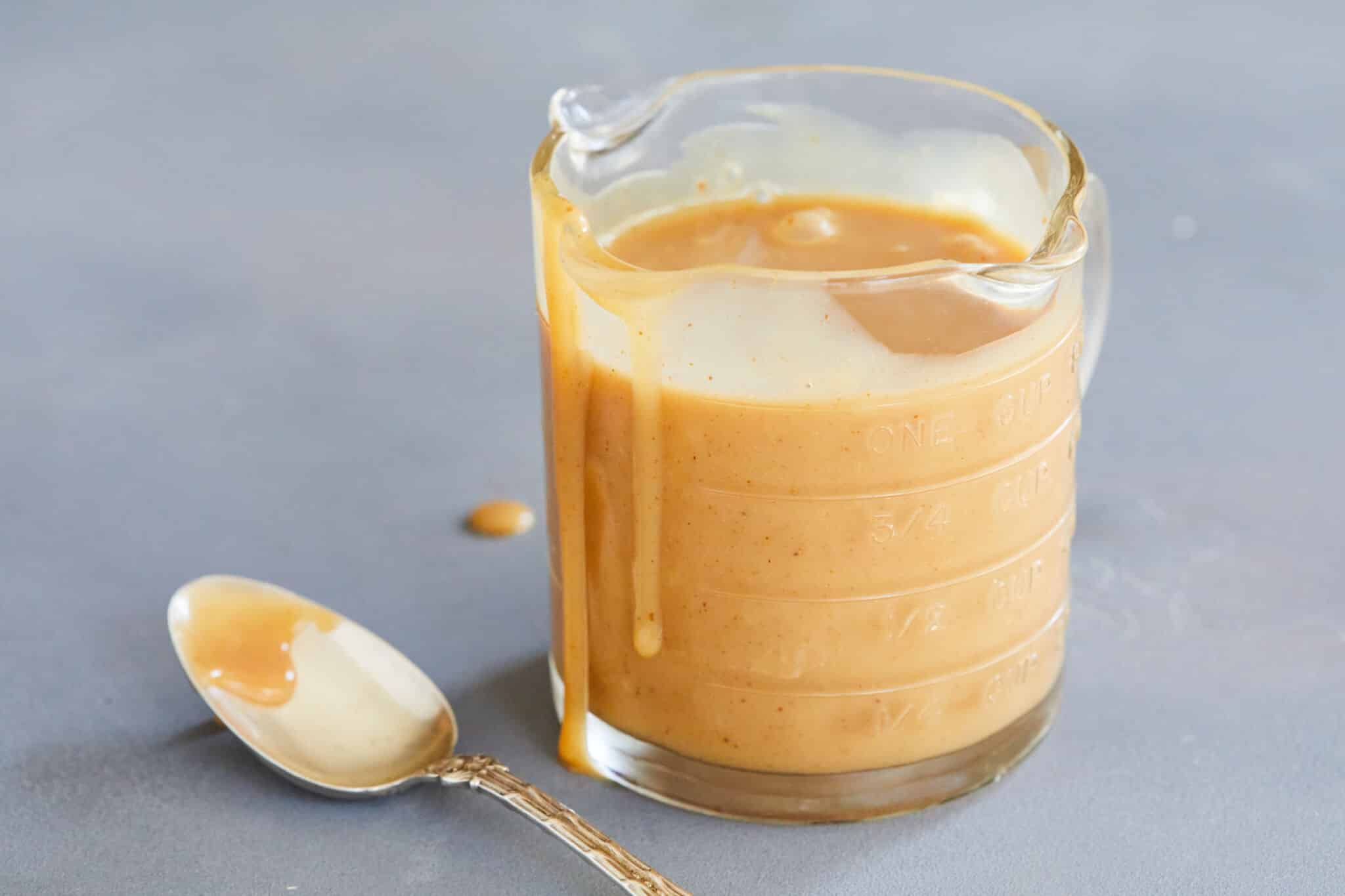 The velvety smooth peanut butter ice cream sauce is served in a 3-spout glass measuring jug, with extra dripping down on the side and a spoon next to it.