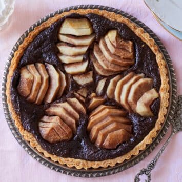 A decadent Pear and Chocolate Frangipane Tart is served on a silver platter. It has golden crispy crust, a lush almond cocoa filling topped with meltingly tender sliced pears.