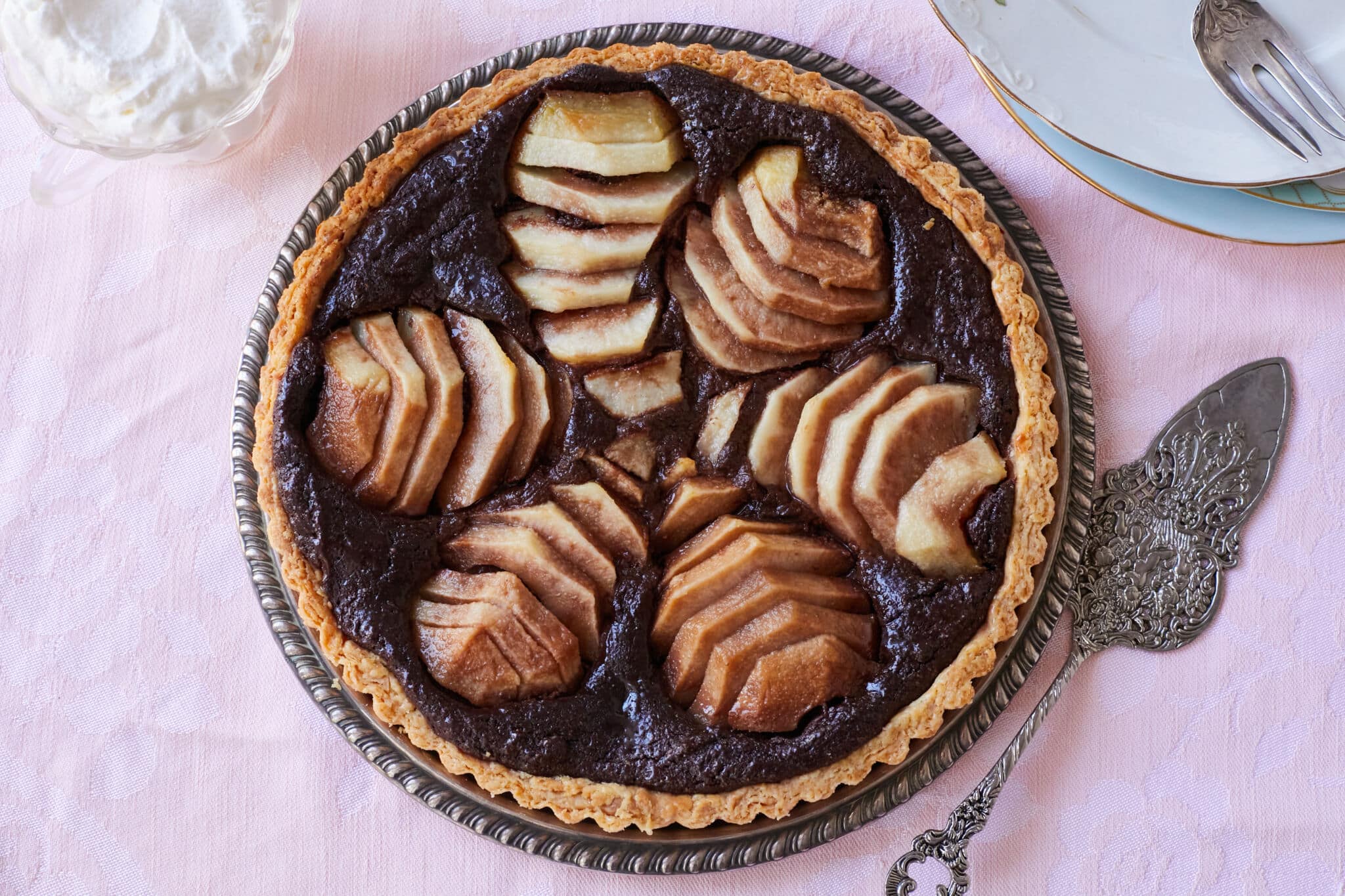 A decadent Pear and Chocolate Frangipane Tart is served on a silver platter. It has golden crispy crust, a lush almond cocoa filling topped with meltingly tender sliced pears.
