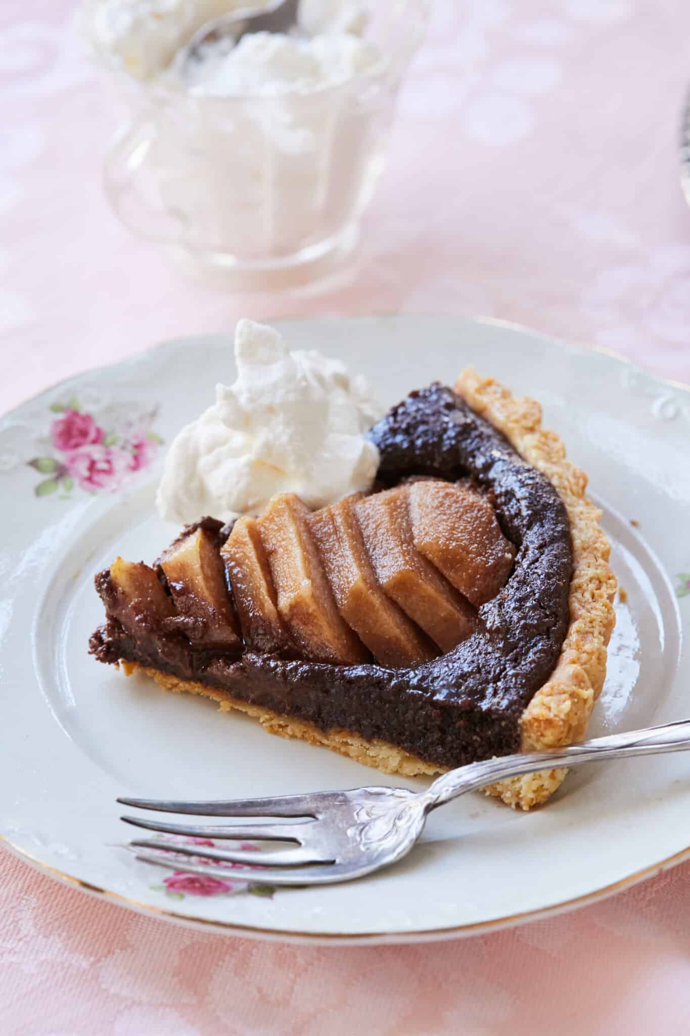 A slice of decadent Pear and Chocolate Frangipane Tart is served on a white porcelain dessert plate with golden trim. The close shot shows the tart has golden crispy crust, a lush almond cocoa filling topped with meltingly tender sliced pears and freshly whipped cream. More whipped cream is in a class cup on the side. 