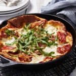 The Dutch Baby is perfectly baked in a cast iron skillet cooling on a black wire rack. The pancake has golden crispy edges, soft silky custard cheese center, toped with savory prosciutto and fresh green arugula. 3 sets of paste and silverware are in the top left corner.