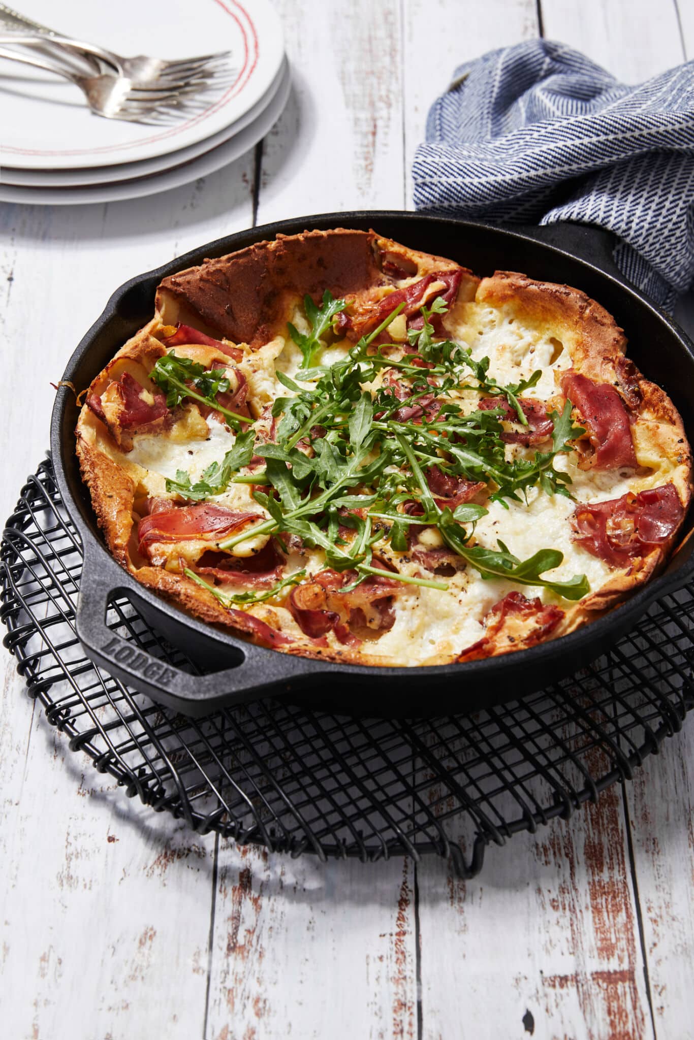 The Dutch Baby is perfectly baked in a cast iron skillet cooling on a black wire rack. The pancake has golden crispy edges, soft silky custard cheese center, toped with savory prosciutto and fresh green arugula. 3 sets of plates and silverware are in the top left corner for serving.