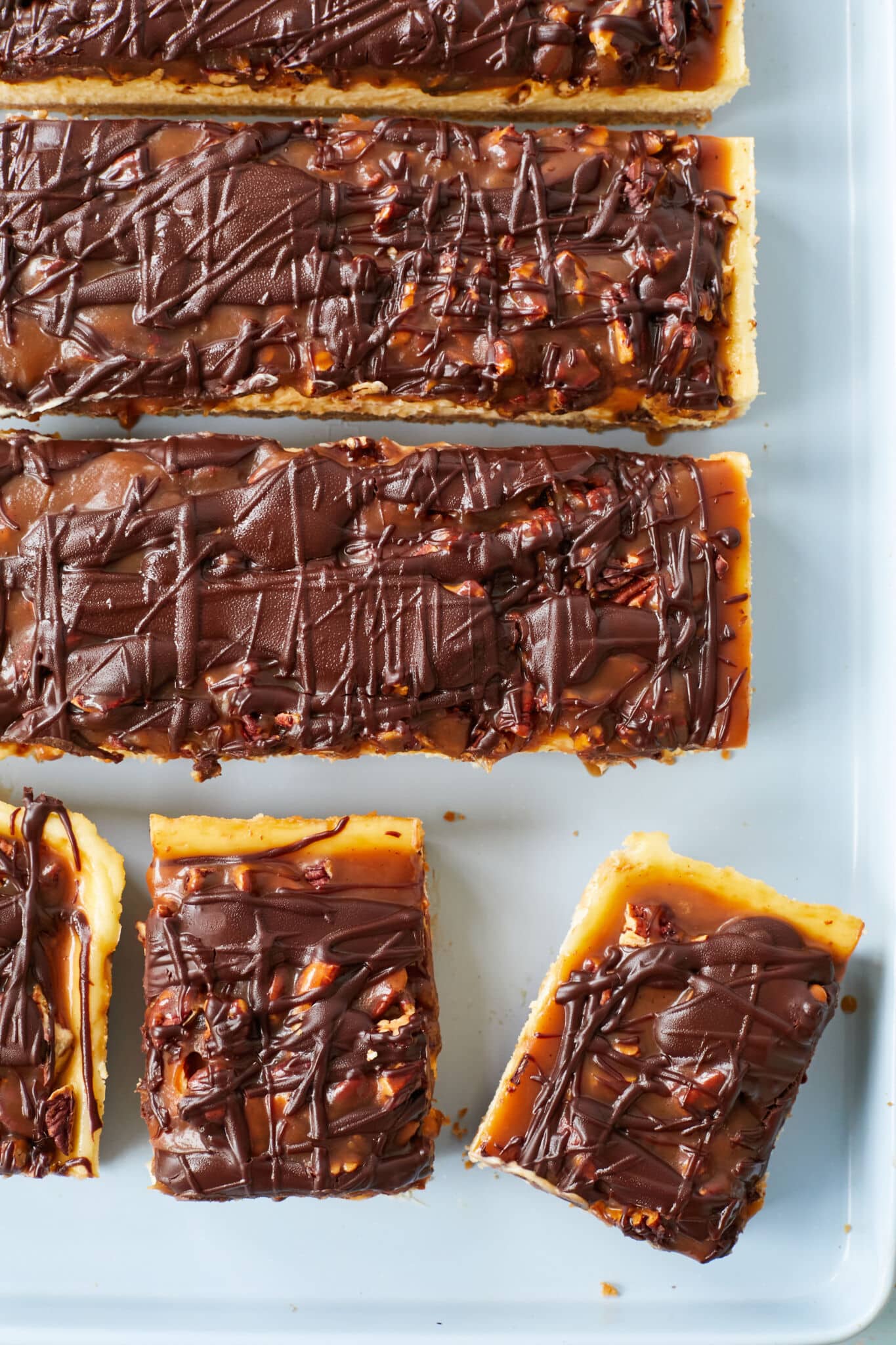Turtle Cheesecake Bars are baked and cut into strips and squares in a light blue baking pan. An overhead shot shows the buttery caramel, nuts, and decadent chocolate ganache.