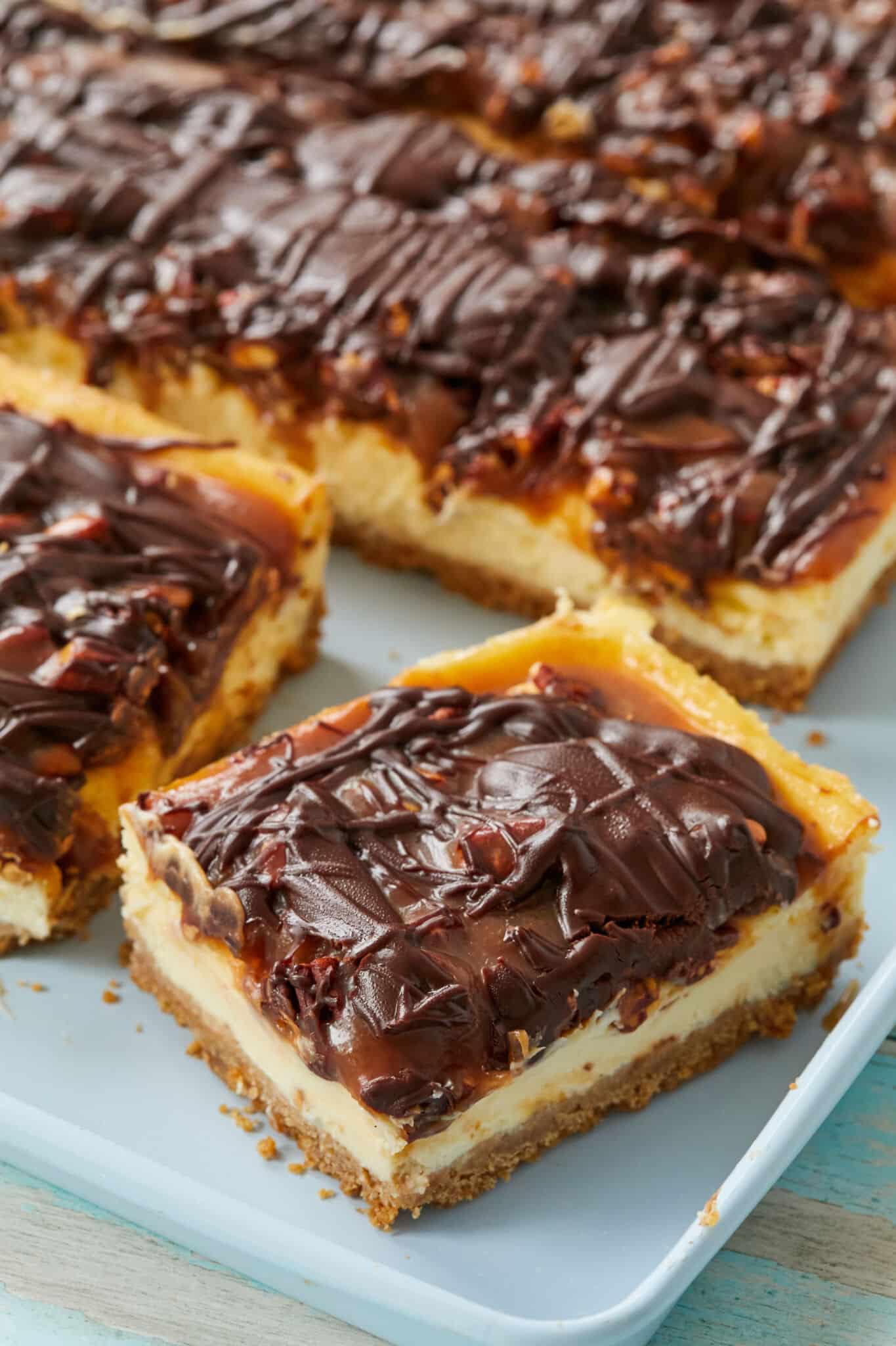 Turtle Cheesecake Bars are baked and cut into squares in a light blue baking pan. They have thick, crumbly graham cracker crust, topped with velvety cheesecake filling covered in buttery caramel, nuts, and drizzled with decadent chocolate ganache.