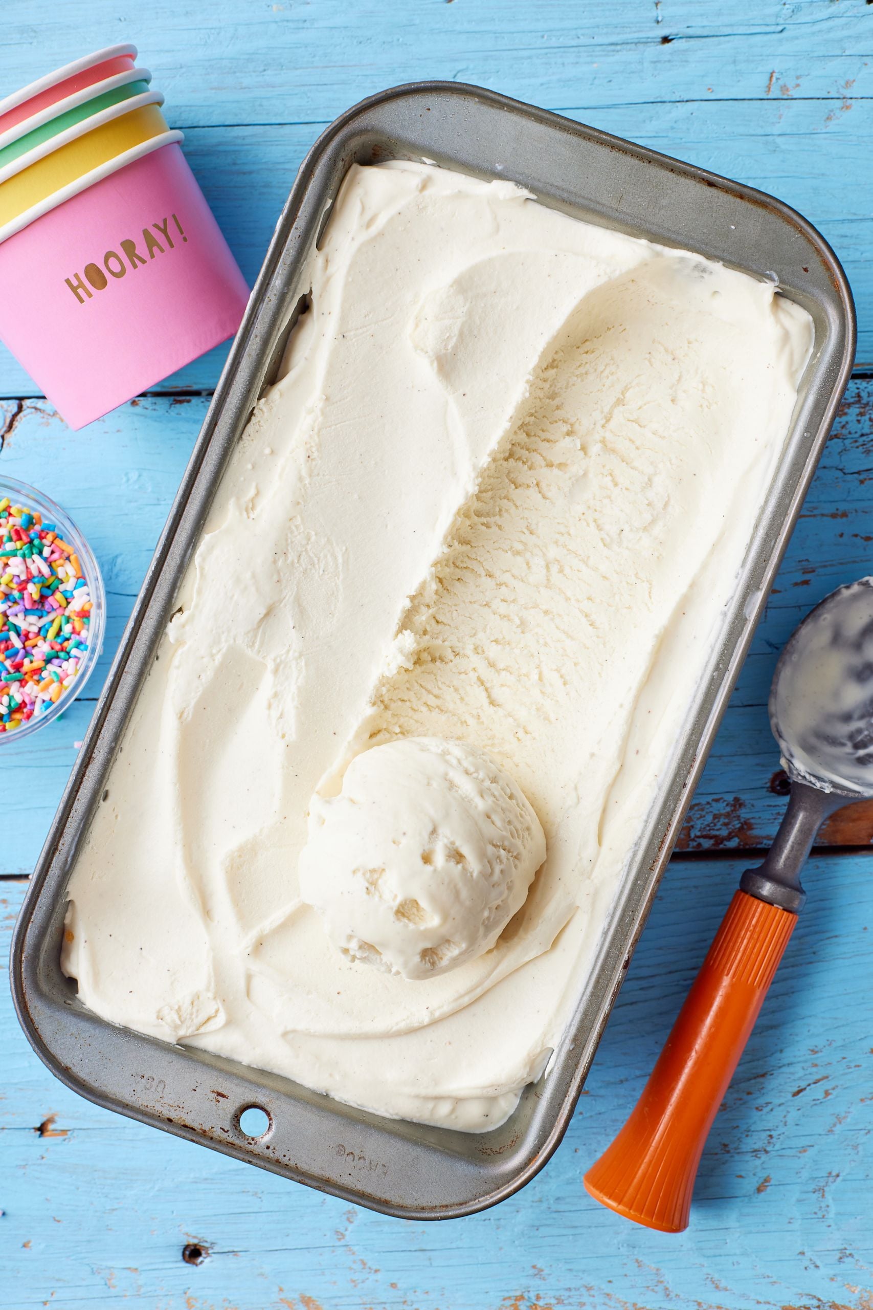 A top-down view of a loaf tin of vanilla ice cream with paper cups and a scoop on the side 