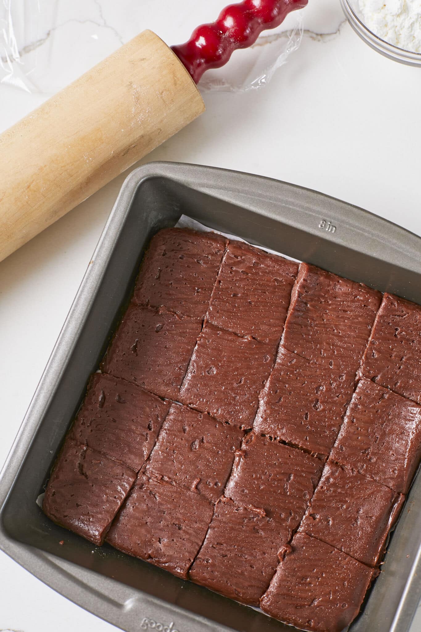 The brown batter was chilled in an 8 by 8 inch metal baking tray and cut into 16 squares. A wooden rolling pin with a red handle and a small glass bowl of cornstarch are placed on the side. 