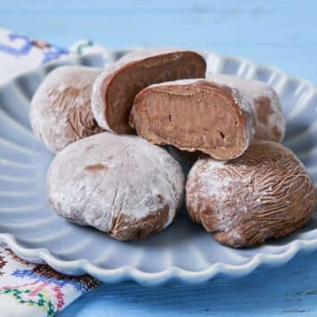 Five Chocolate Mochi Ice Cream balls are served on a blue scalloped plate with the top one cut into half. The exterior mochi is dark brown soft and chewy traditional Japanese sticky rice cake. The inside is glossy velvety, cold ice cream.