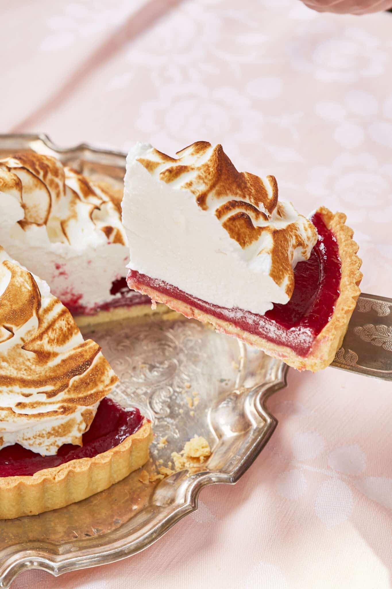 A slice of Mile-High Raspberry Meringue Pie is cut and being lifted from the silver serving plate. The side of the slice shows its flaky crust, creamy tangy sweet raspberry curd filling and silky meringue toasted on top. 