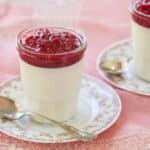 The white silky smooth buttermilk Panna Cotta is topped with glossy vibrant red raspberry jam, served in a mason jar on a floral plate with a vintage dessert spoon.