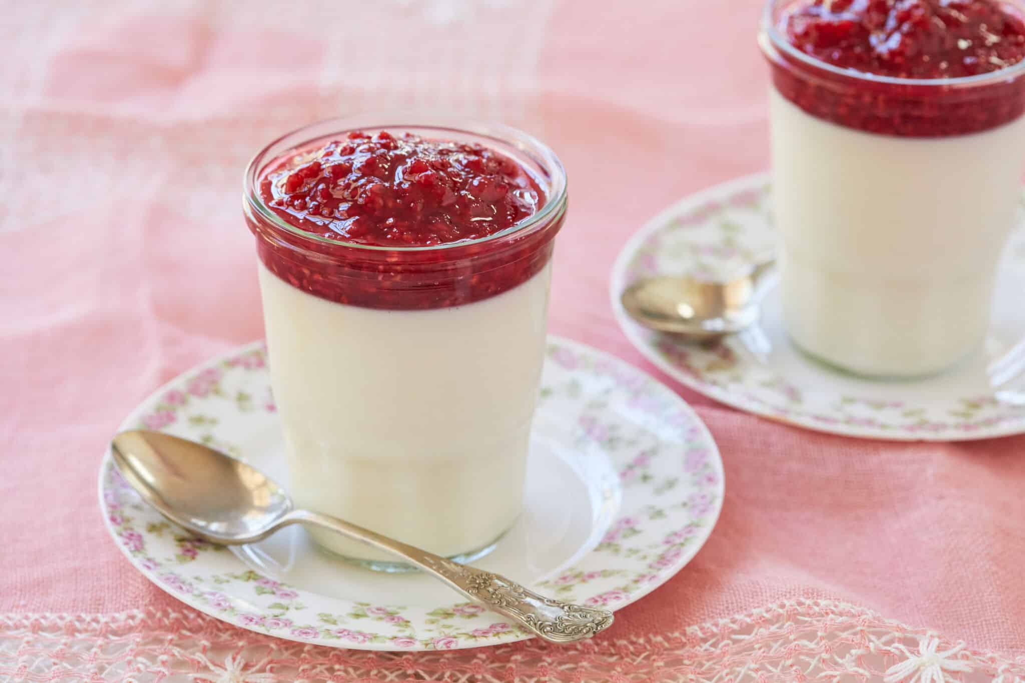 The white silky smooth buttermilk Panna Cotta is topped with glossy vibrant red raspberry jam, served in a mason jar on a floral plate with a vintage dessert spoon.