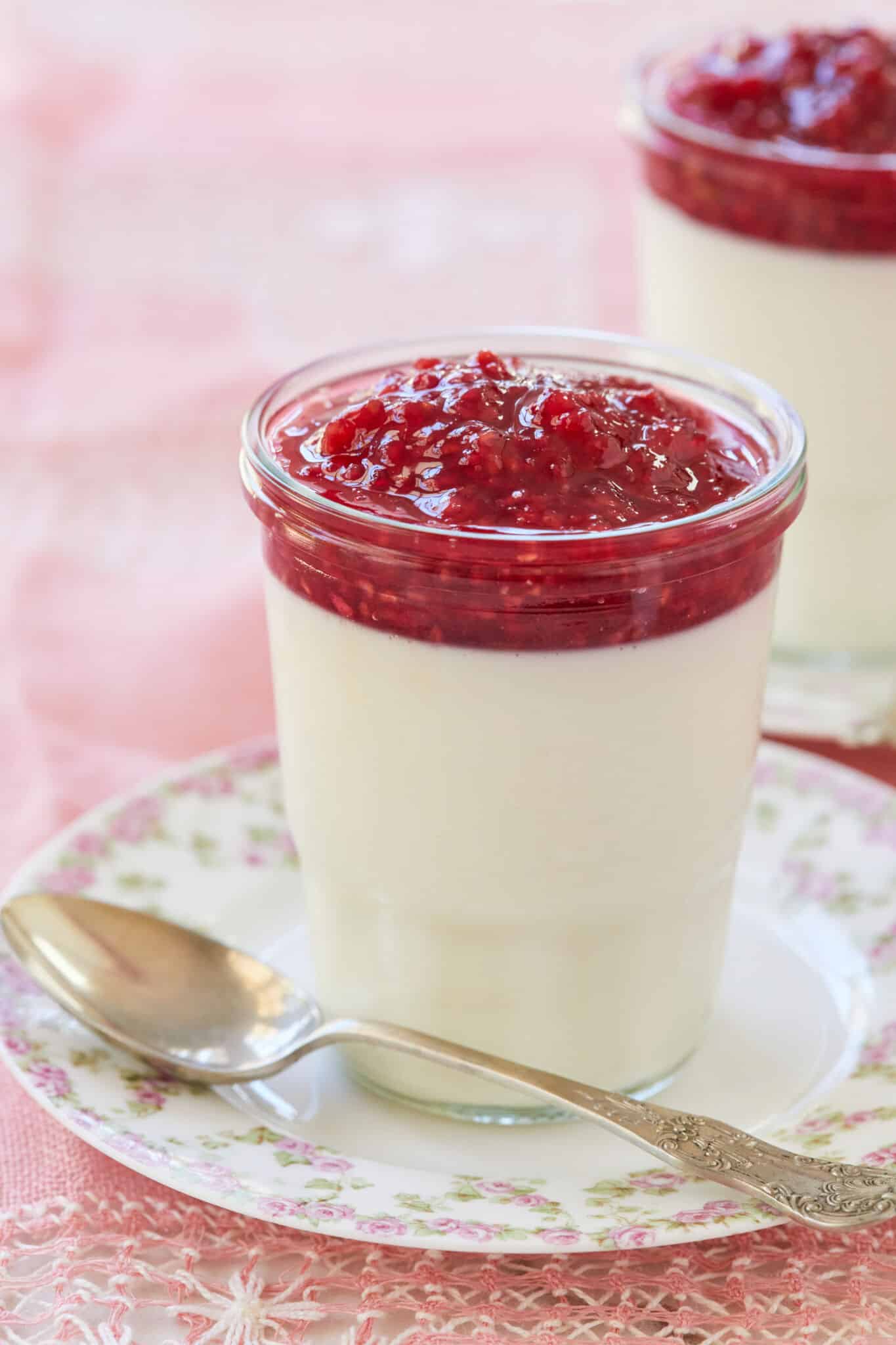 The white silky smooth buttermilk Panna Cotta is topped with glossy vibrant red raspberry jam, served in a mason jar on a floral plate with a vintage dessert spoon. 
